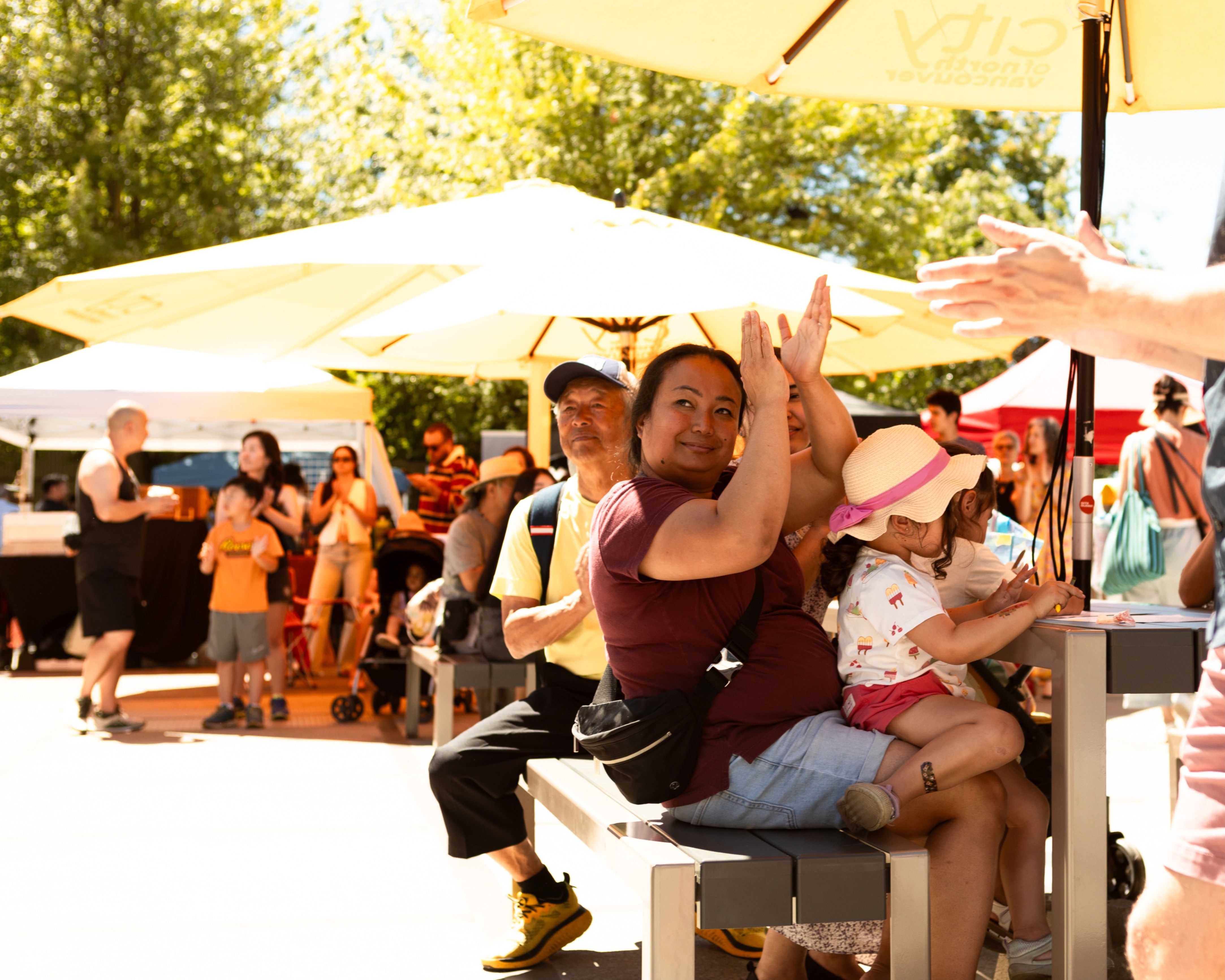 A person clapping hands while sitting on a bench with a child wearing a hat, surrounded by people at an outdoor event with umbrellas.