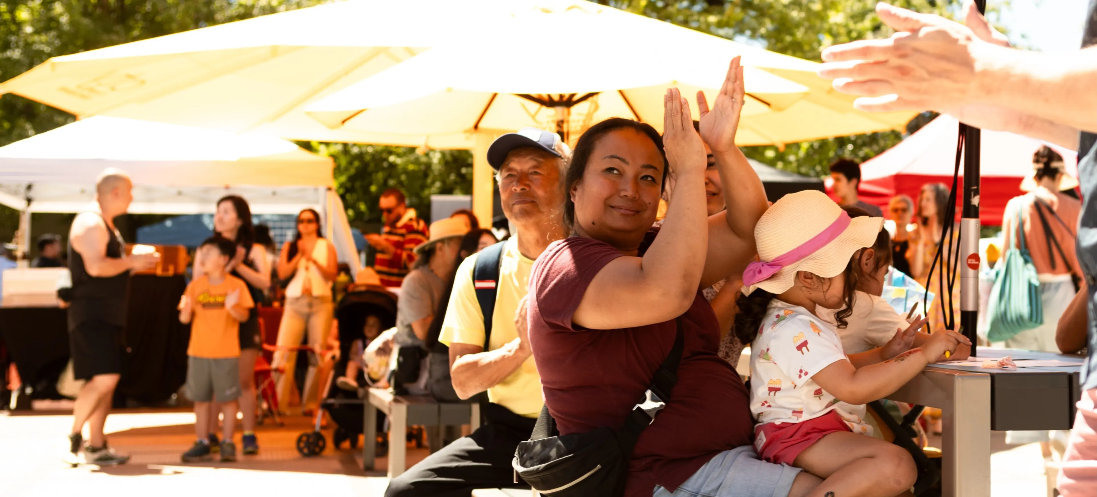 A person clapping hands while sitting on a bench with a child wearing a hat, surrounded by people at an outdoor event with umbrellas.
