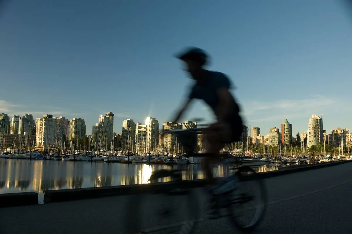 A cyclist bikes past the camera along the Stanley Park Seawall with the Vancouver skyline in the background.