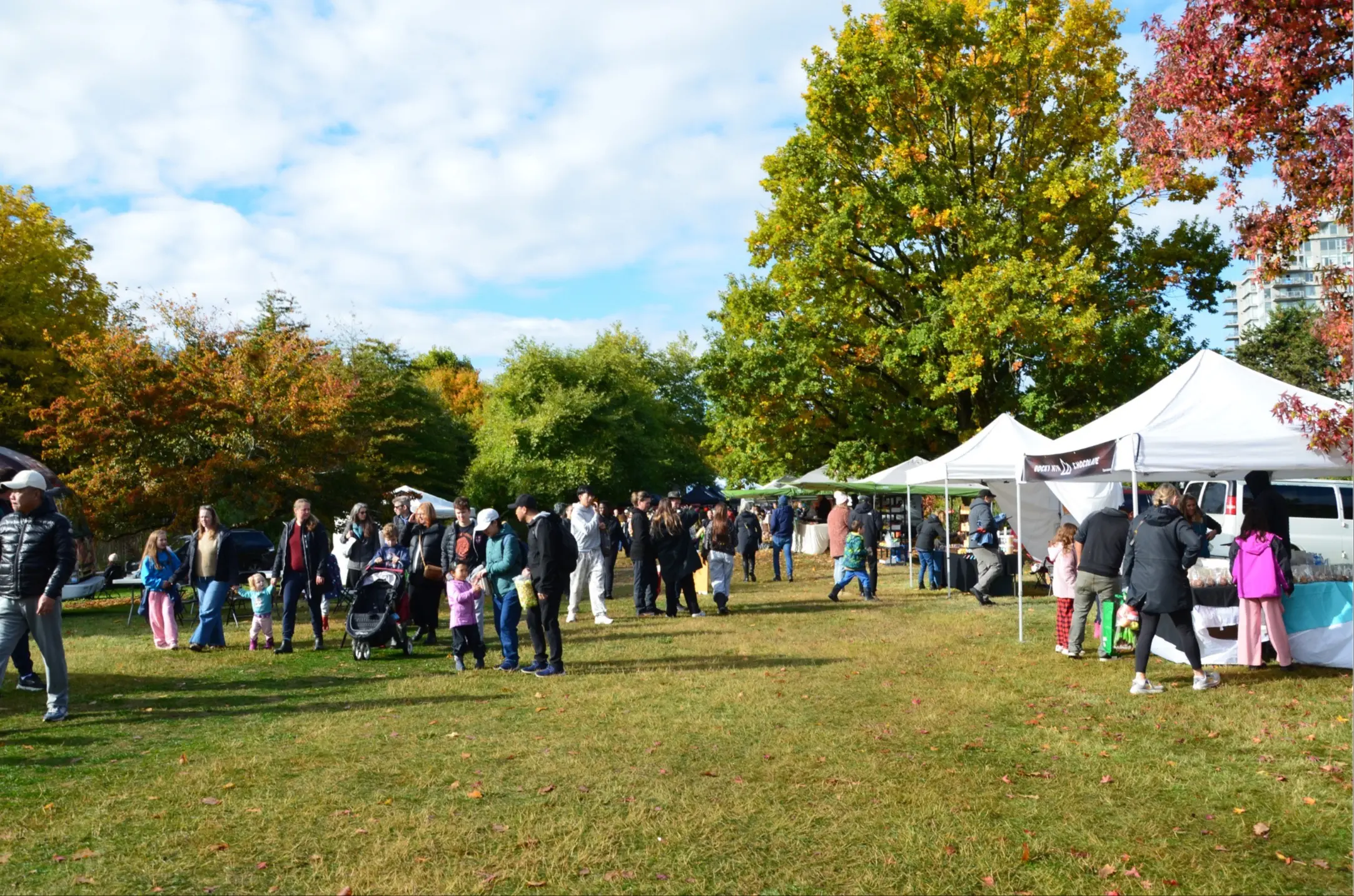 People walking and standing near white tents at an outdoor market on a grassy area with trees showing autumn colors.