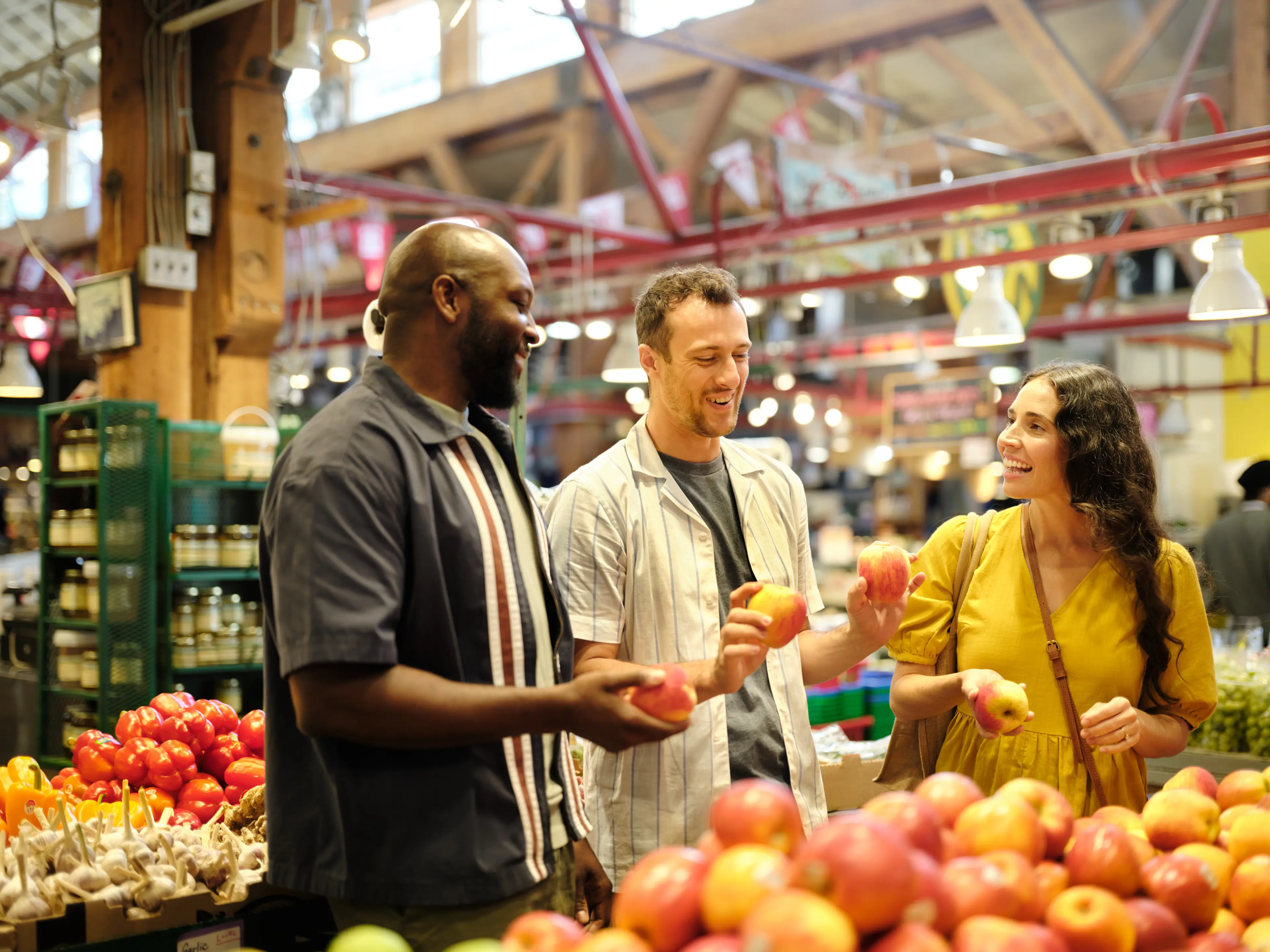 Three people smiling and holding apples while shopping at an indoor market with fruits and vegetables around.