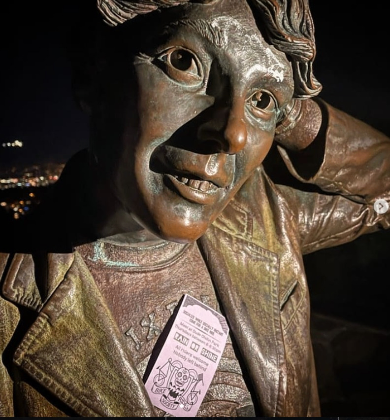 Bronze statue of a person with a hand behind their head, wearing a jacket and holding a pink flyer with text and illustrations.