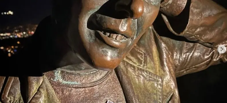 Bronze statue of a person with a hand behind their head, wearing a jacket and holding a pink flyer with text and illustrations.