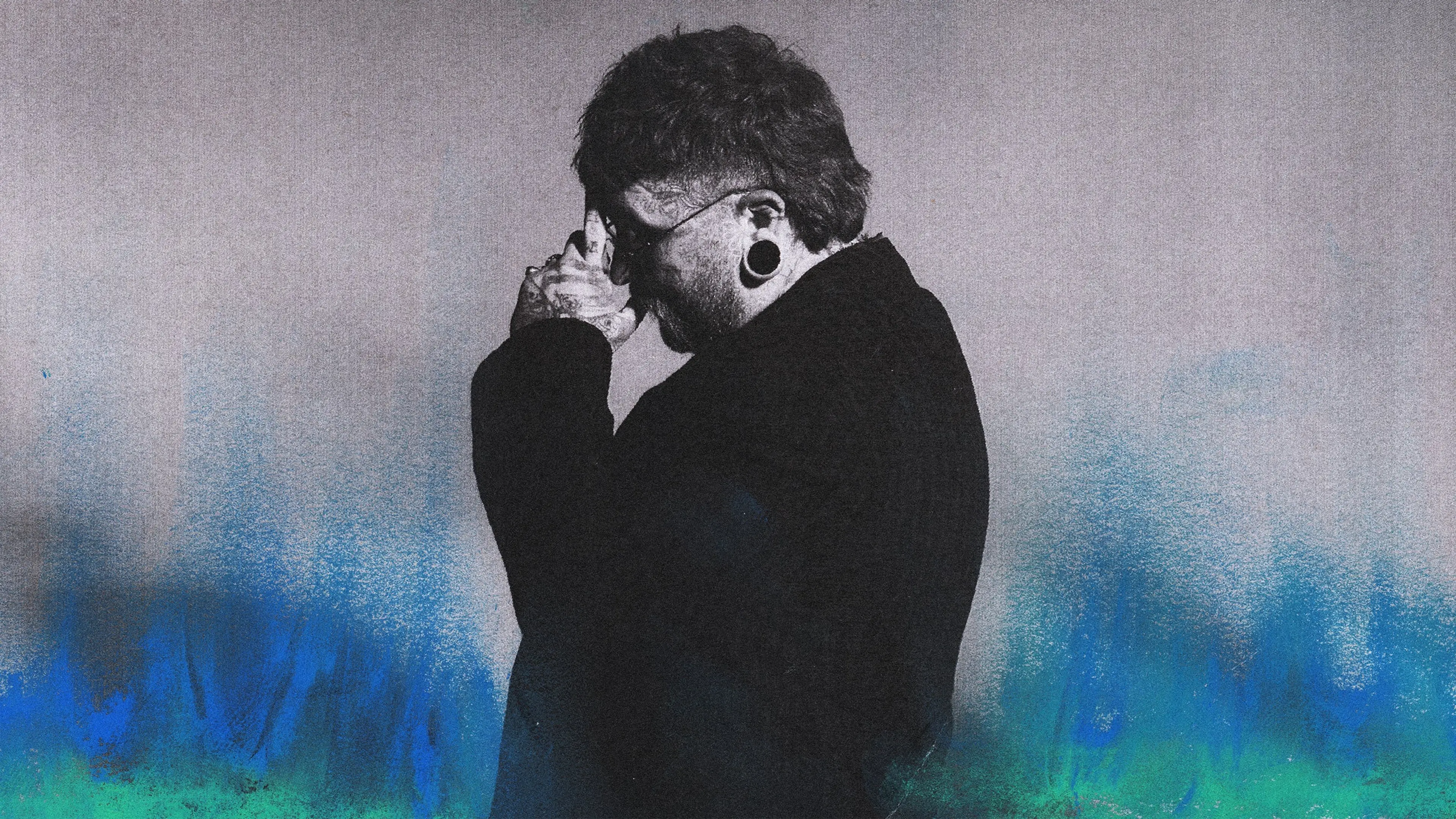 Person with stretched ear piercings and tattoos holding hands near face, standing against a gray and blue gradient background.