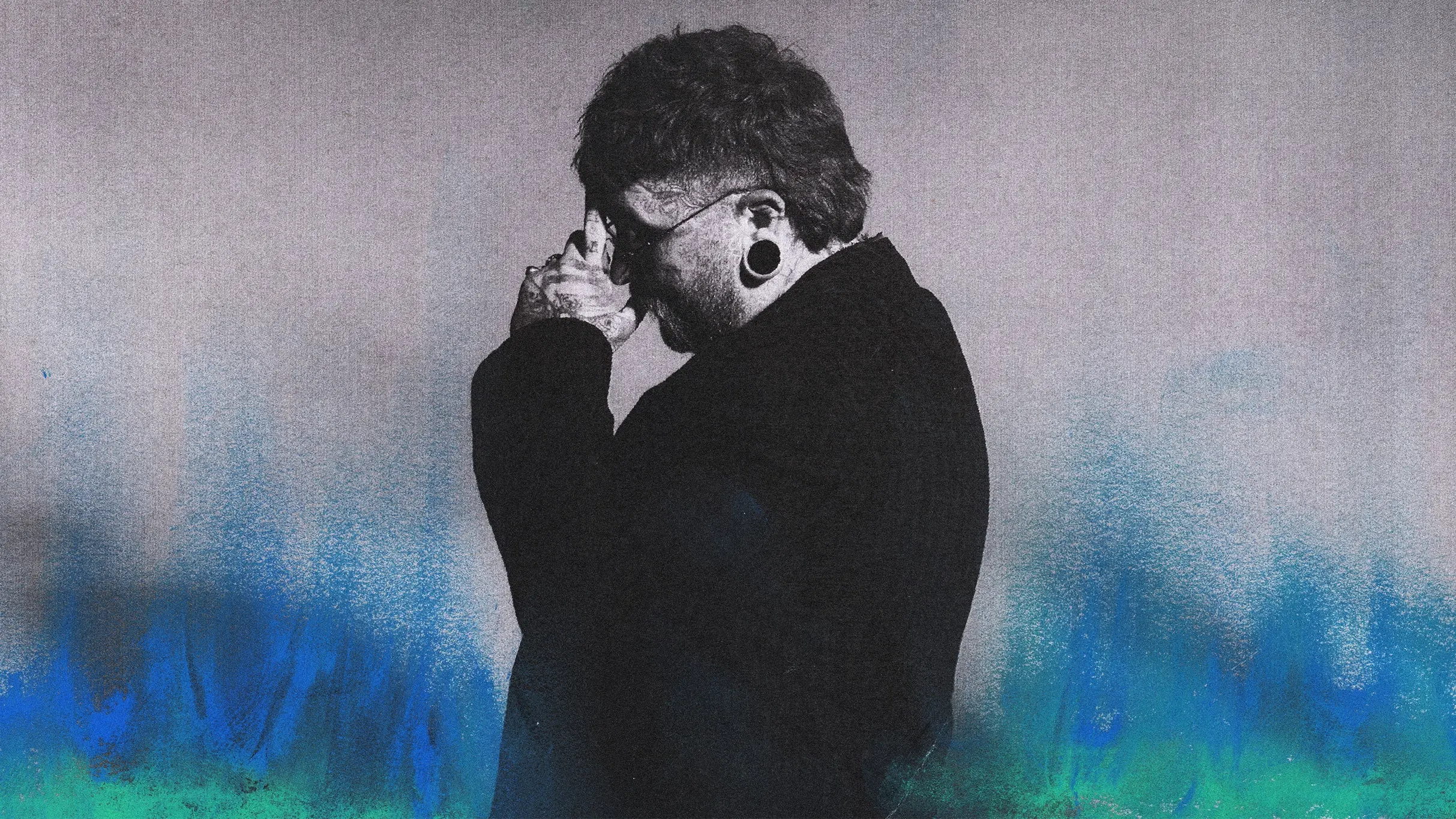 Person with stretched ear piercings and tattoos holding hands near face, standing against a gray and blue gradient background.