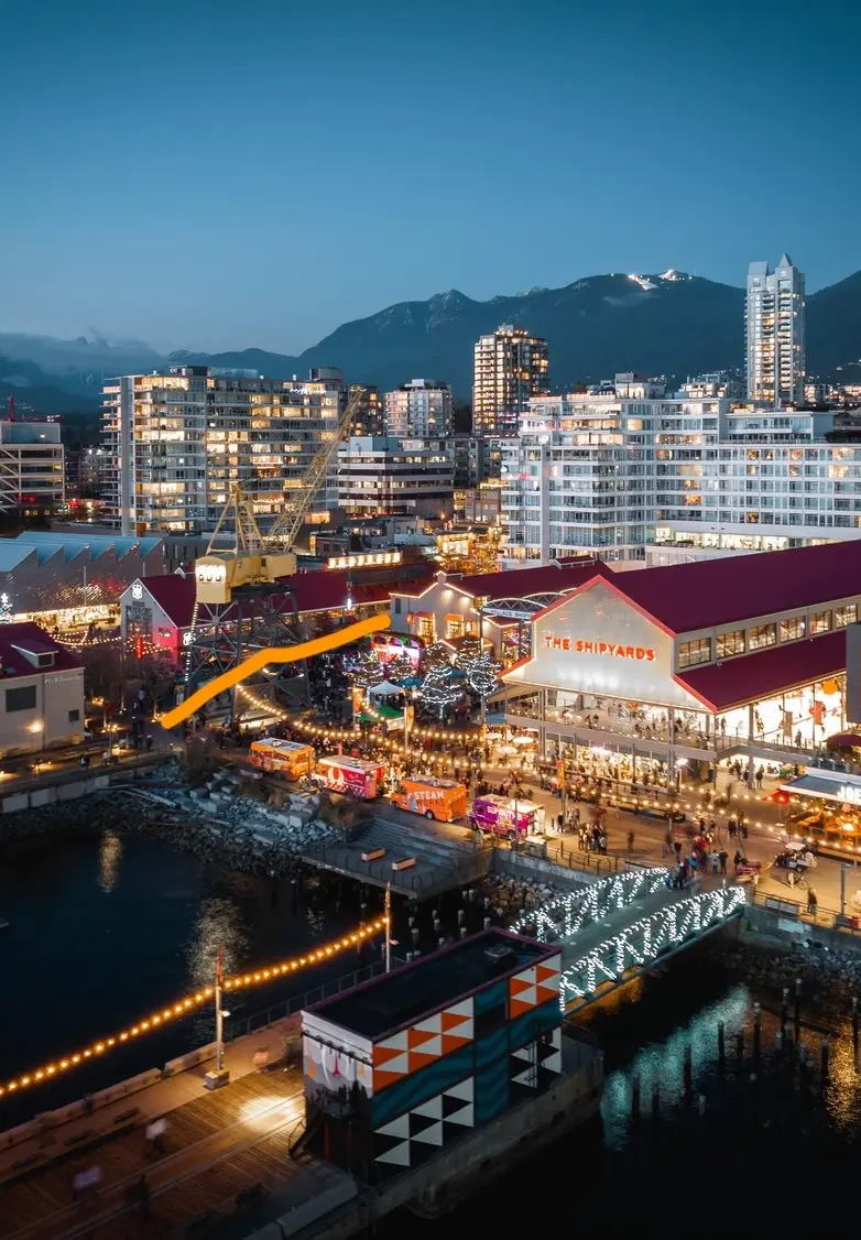 Evening view of a waterfront area with lit buildings, a bridge decorated with lights, and mountains in the background.