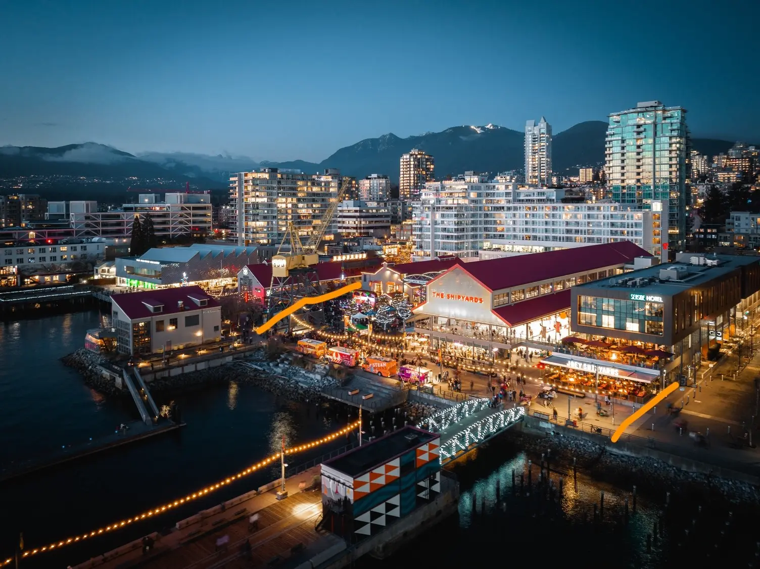 Evening view of a waterfront area with lit buildings, a bridge decorated with lights, and mountains in the background.