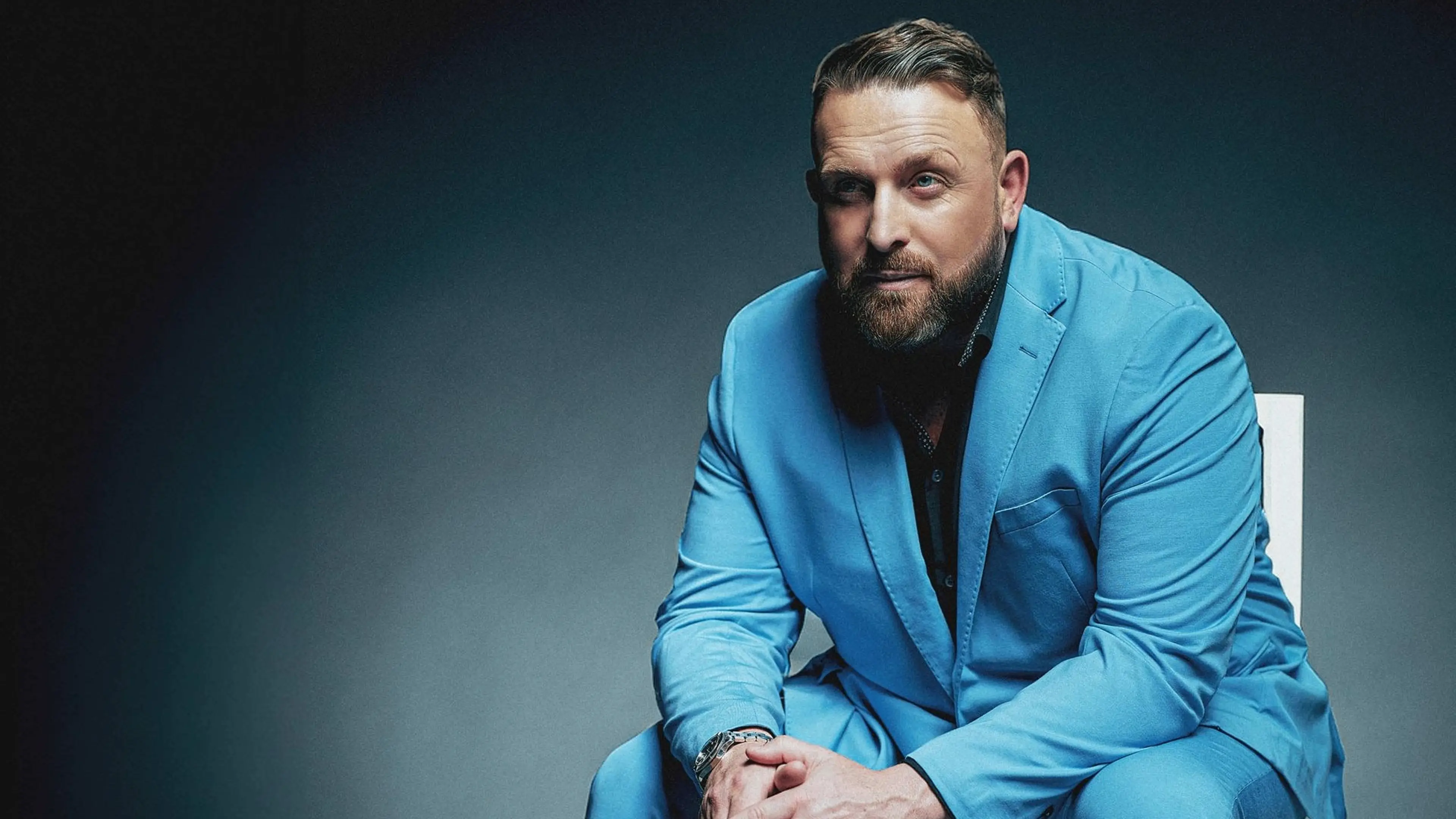 Person with beard wearing a blue suit sitting on a white chair against a dark background.
