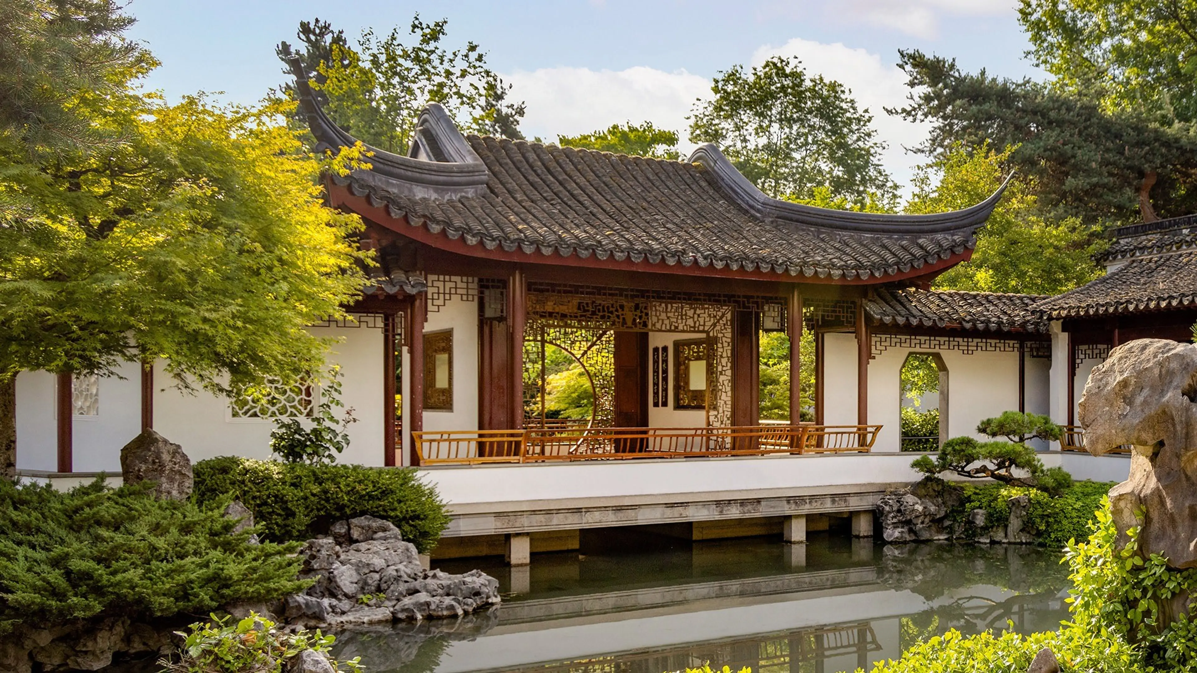 Traditional Chinese garden pavilion with curved roof over a pond surrounded by green trees and rocks.