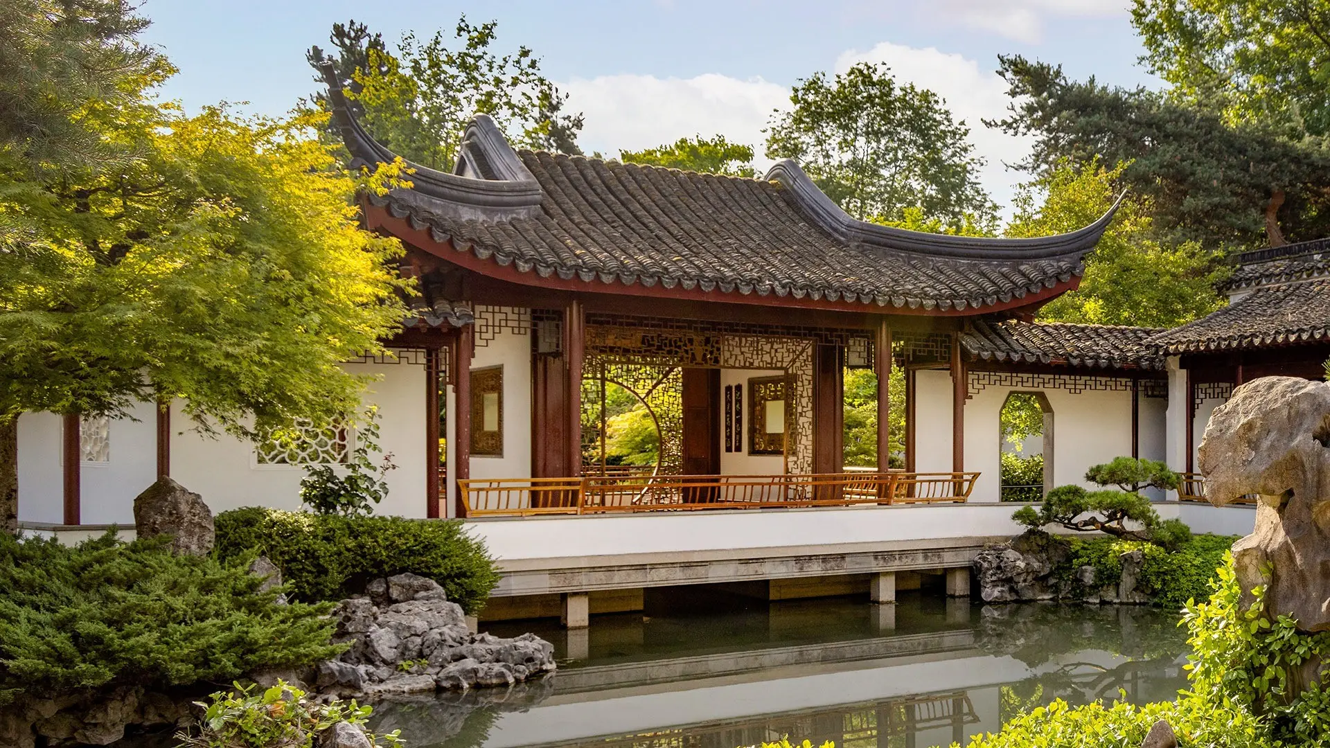 Traditional Chinese garden pavilion with curved roof over a pond surrounded by green trees and rocks.