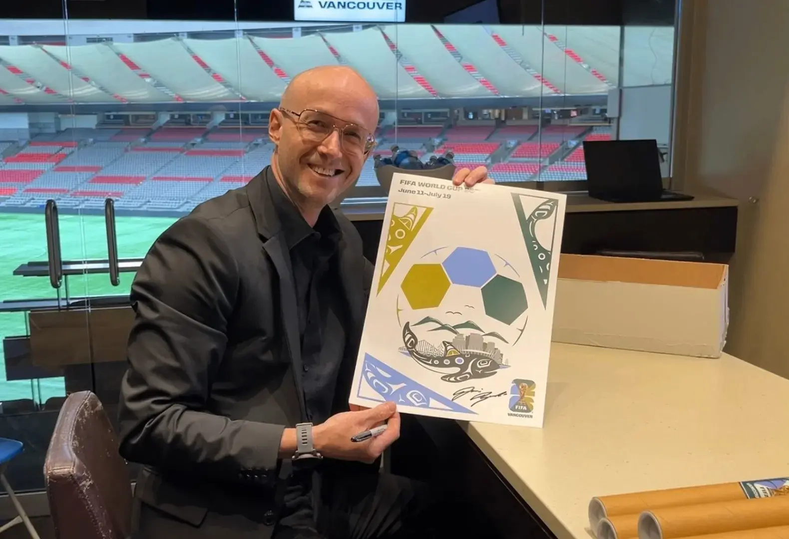 Person in black suit and glasses smiling while holding a signed FIFA World Cup poster inside a stadium suite.