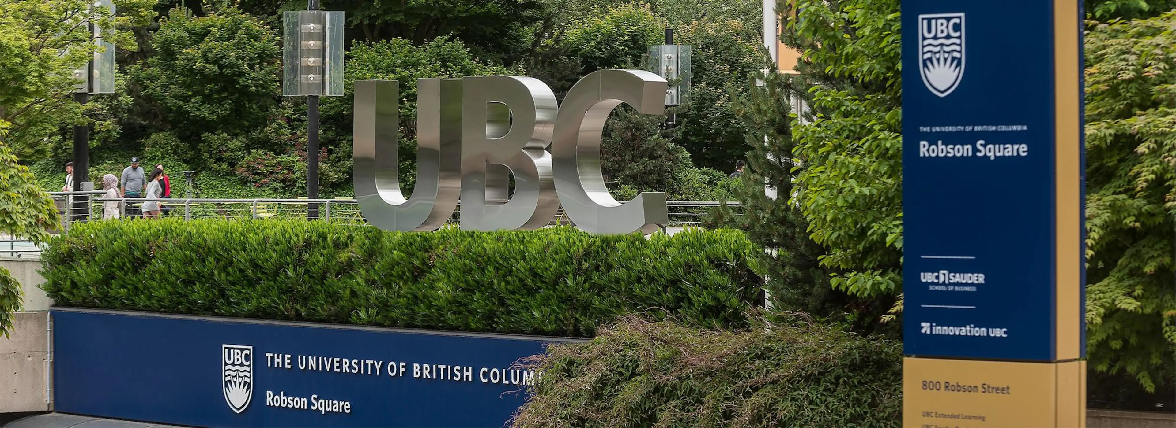 Large metallic UBC letters and signs for University of British Columbia Robson Square surrounded by greenery and people walking in the background.