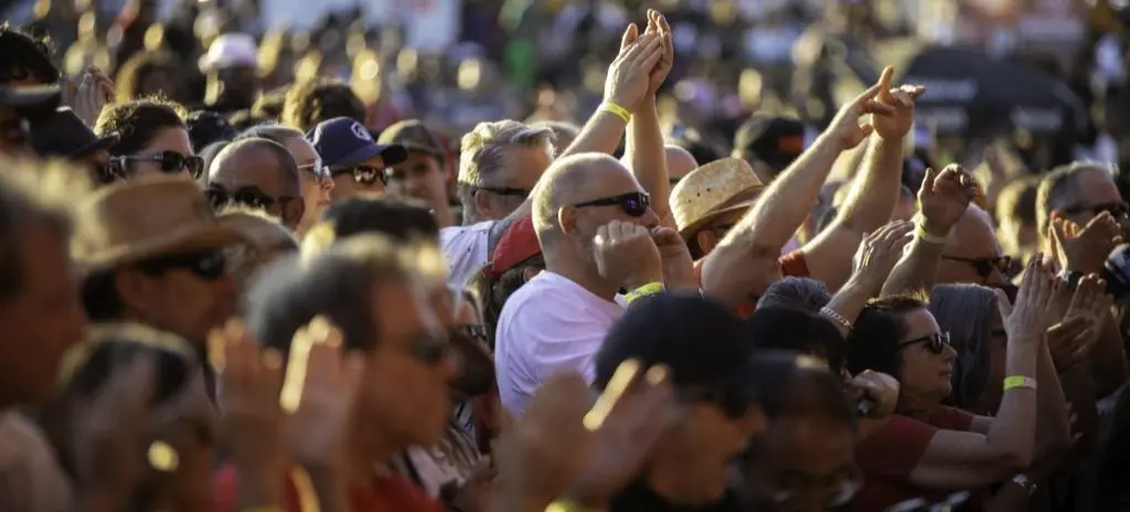 Crowd of people wearing sunglasses and wristbands, raising hands and clapping at an outdoor event.