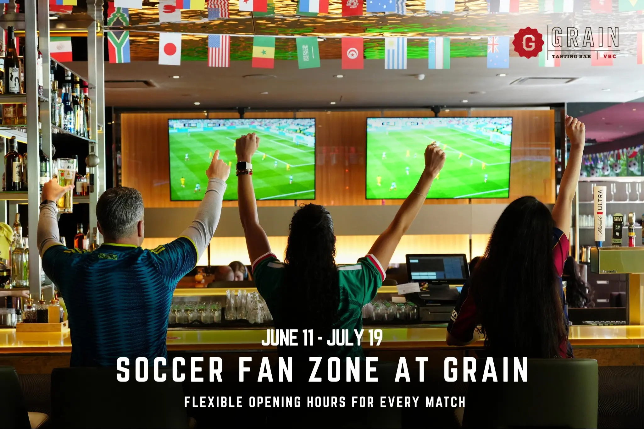 Three people raising arms cheering while watching soccer on two TV screens in a bar decorated with international flags.