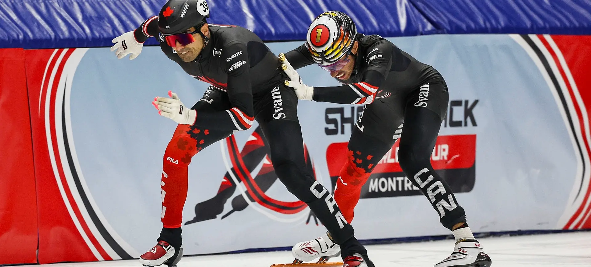 Two speed skaters in black and red suits racing on ice, one pushing the other from behind near a barrier with a logo.