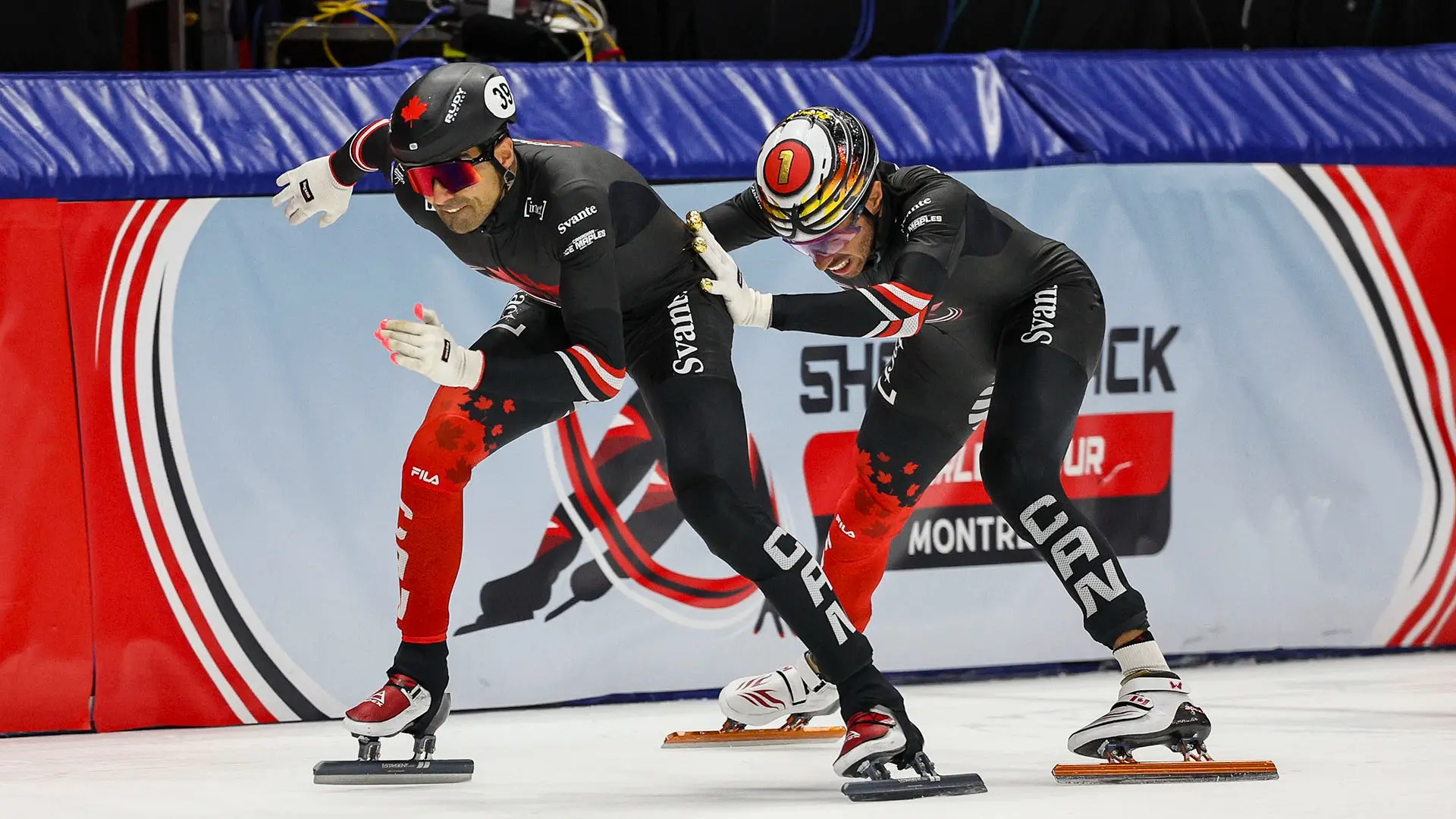 Two speed skaters in black and red suits racing on ice, one pushing the other from behind near a barrier with a logo.
