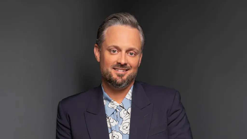 Person with short gray hair and beard wearing a dark blazer and blue floral shirt, smiling against a dark background.