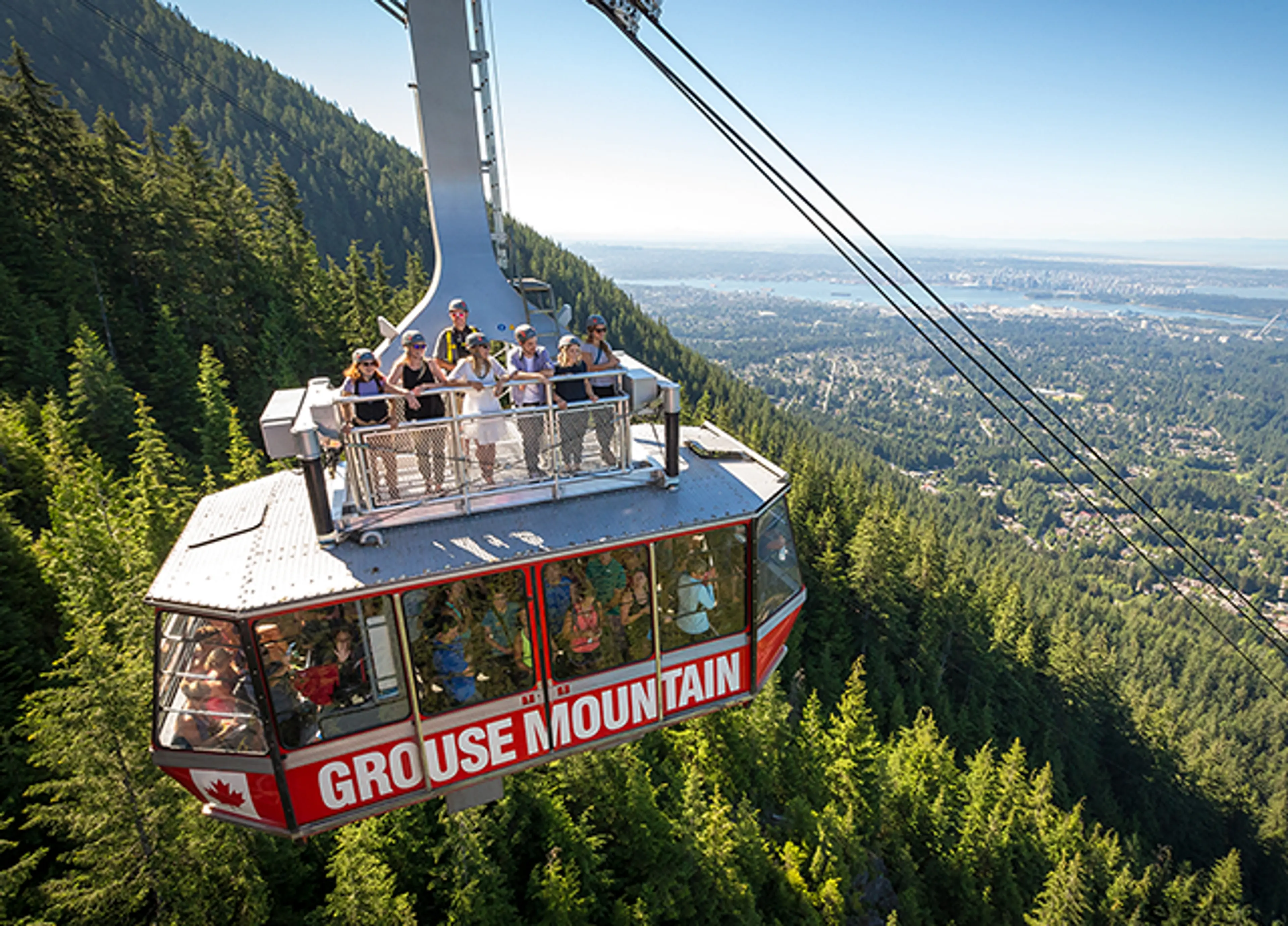 Cable car labeled Grouse Mountain carrying people above a forested mountain with a city and water in the background.
