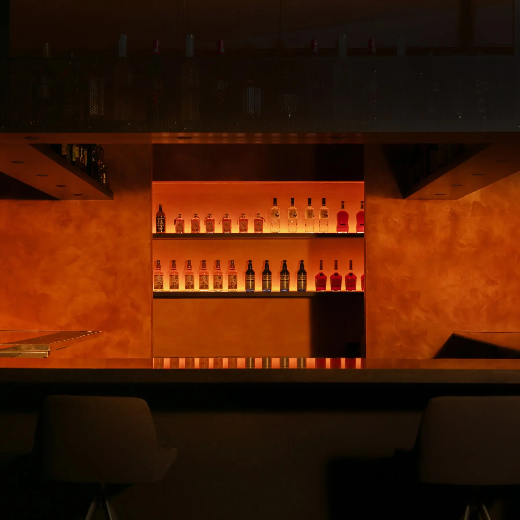 Dimly lit bar with two shelves of various liquor bottles backlit in warm orange light, with two bar stools in front.