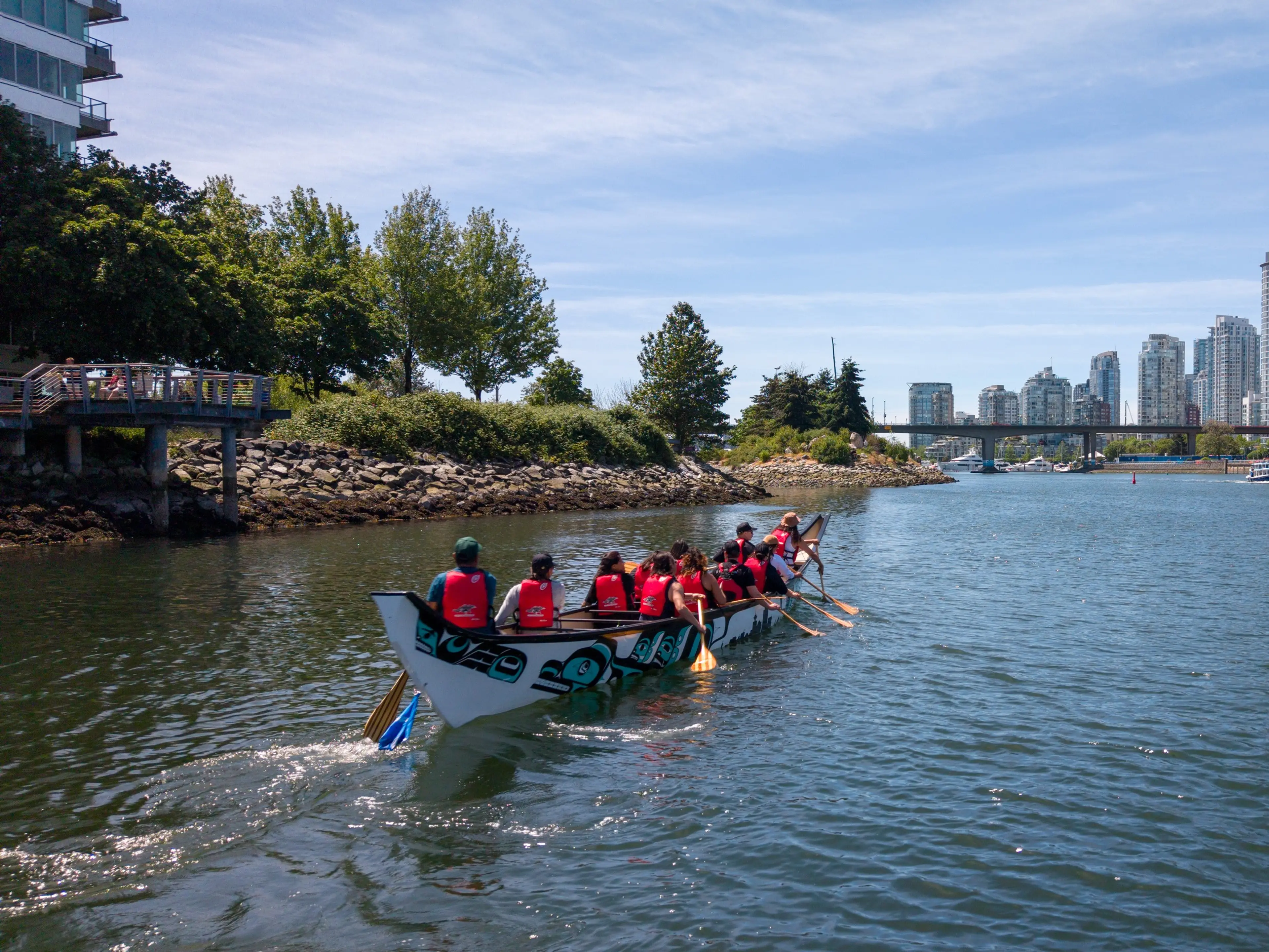 A group of people paddling a decorated canoe on a river with trees and buildings in the background under a blue sky.