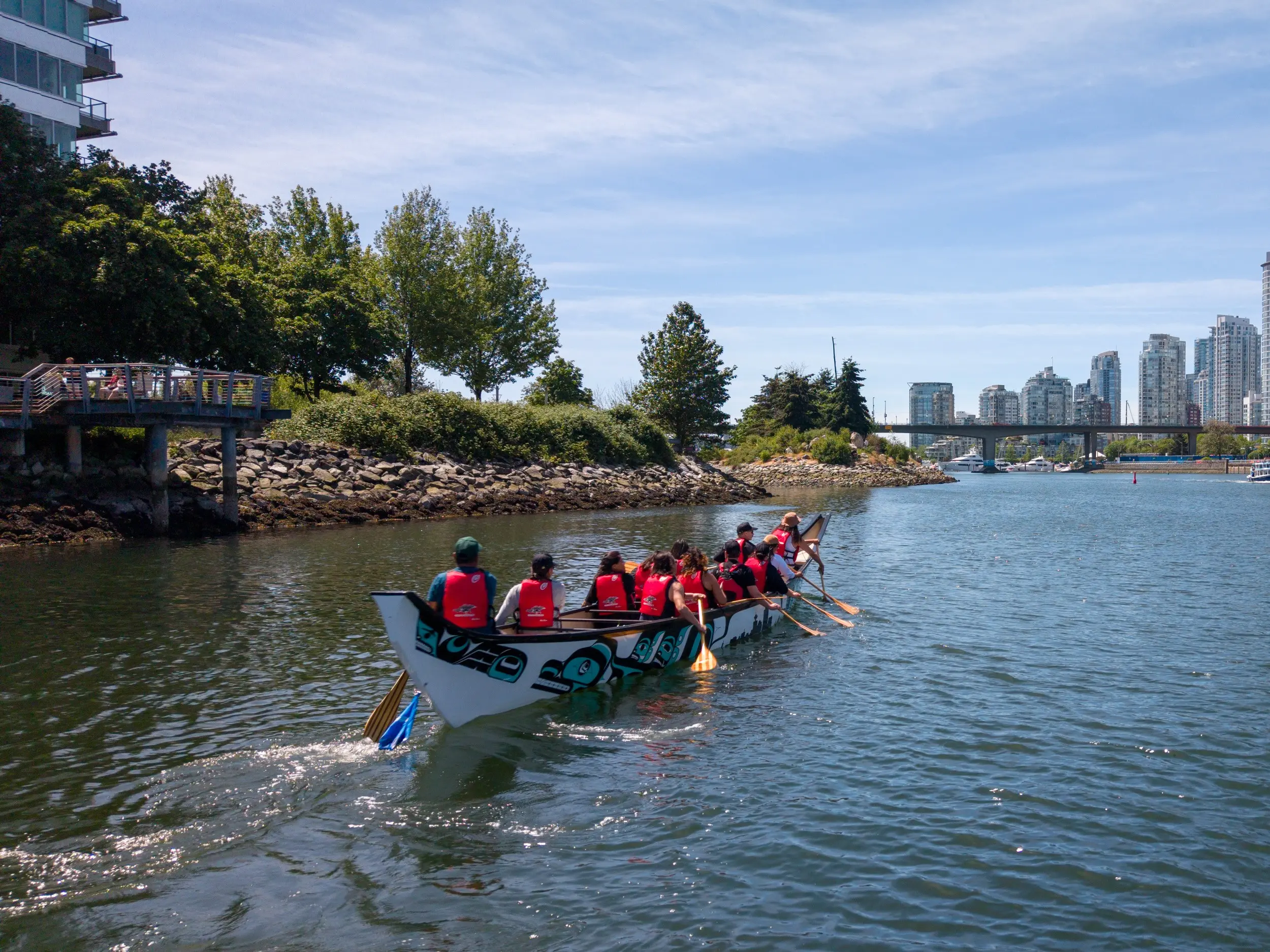 A group of people paddling a decorated canoe on a river with trees and buildings in the background under a blue sky.