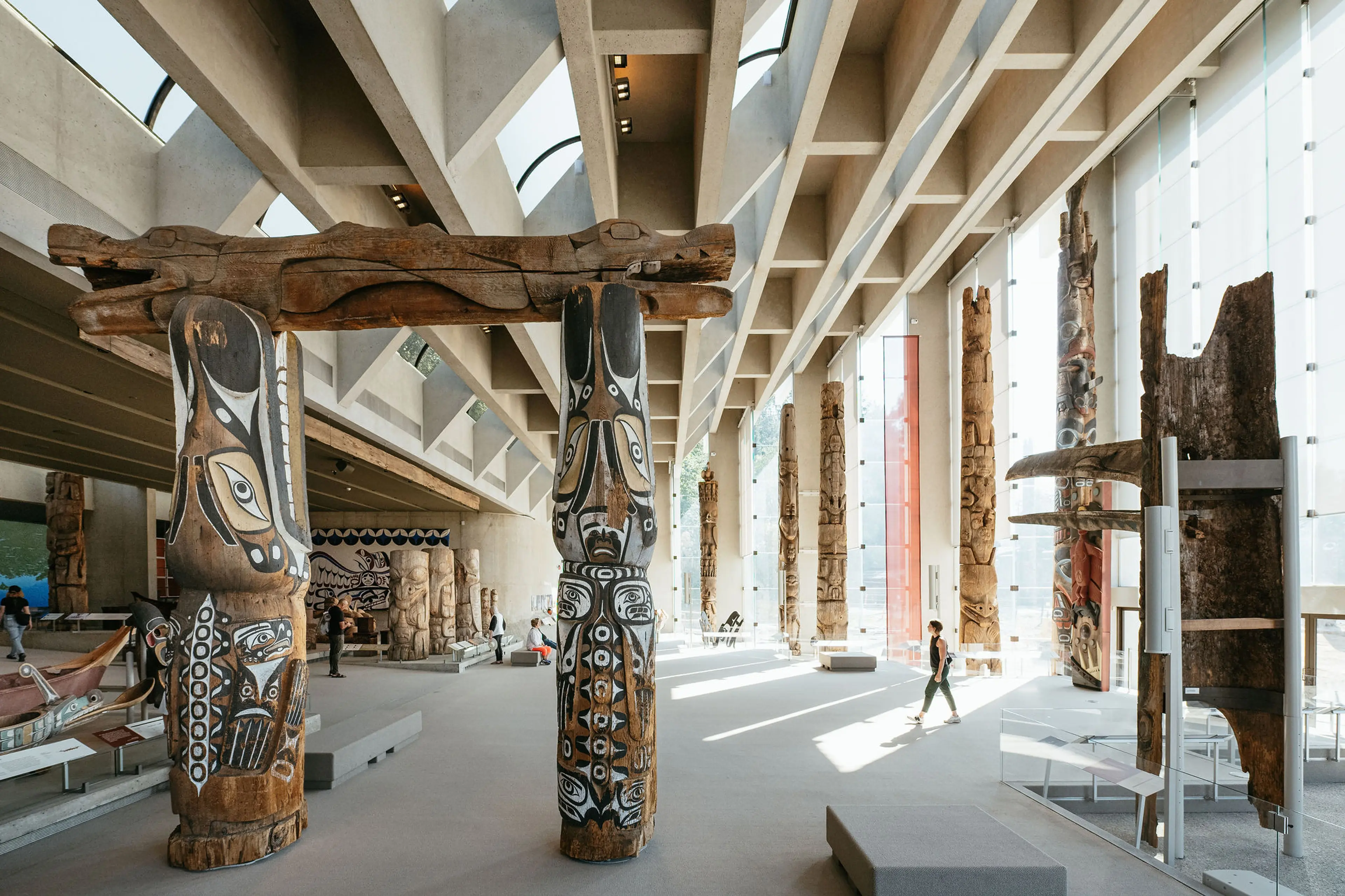 Spacious museum hall with tall wooden totem poles and large windows letting in natural light, a person walking inside.