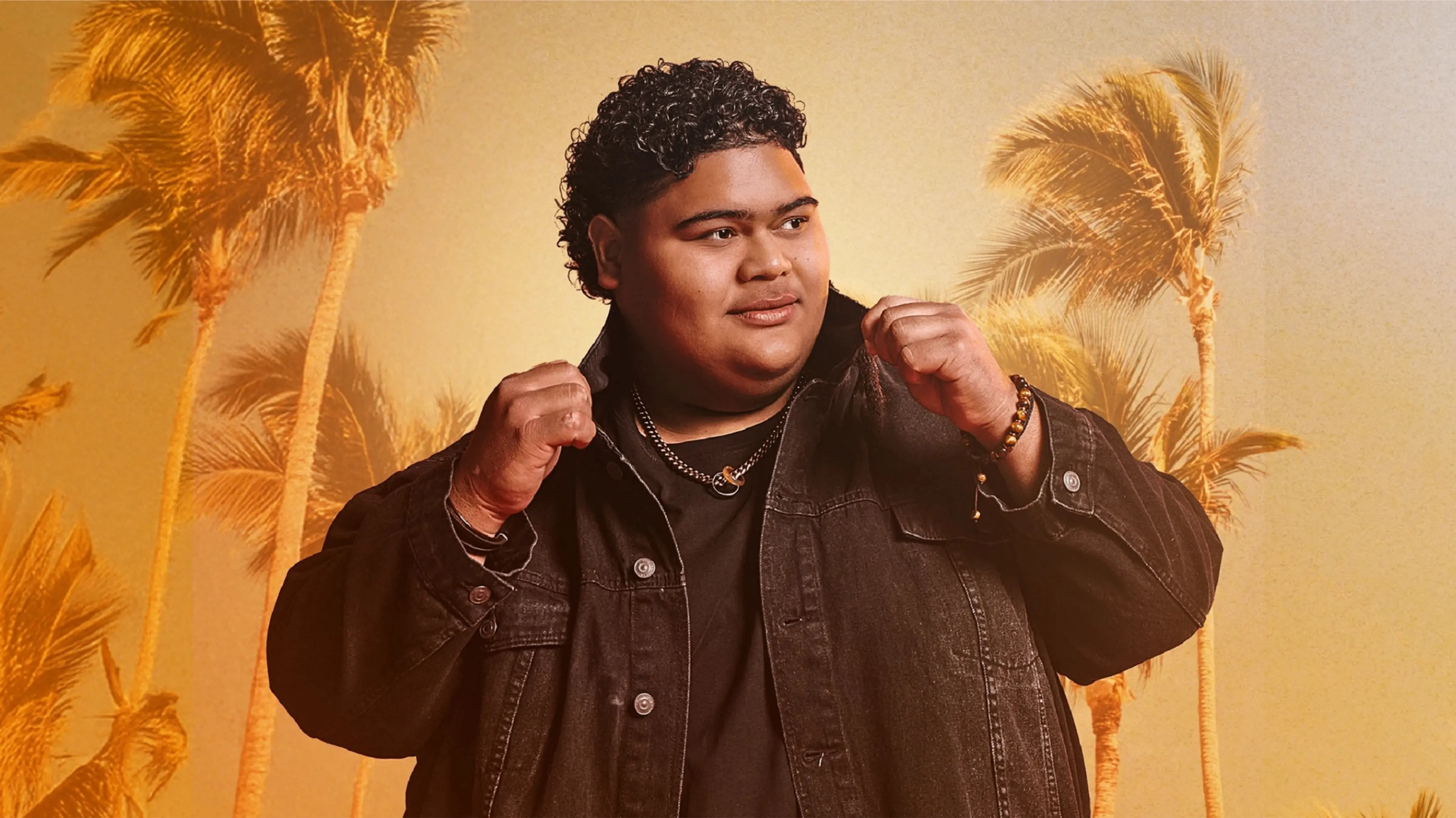 Person with curly hair wearing a black jacket and necklace, posing with fists raised against a background of palm trees.