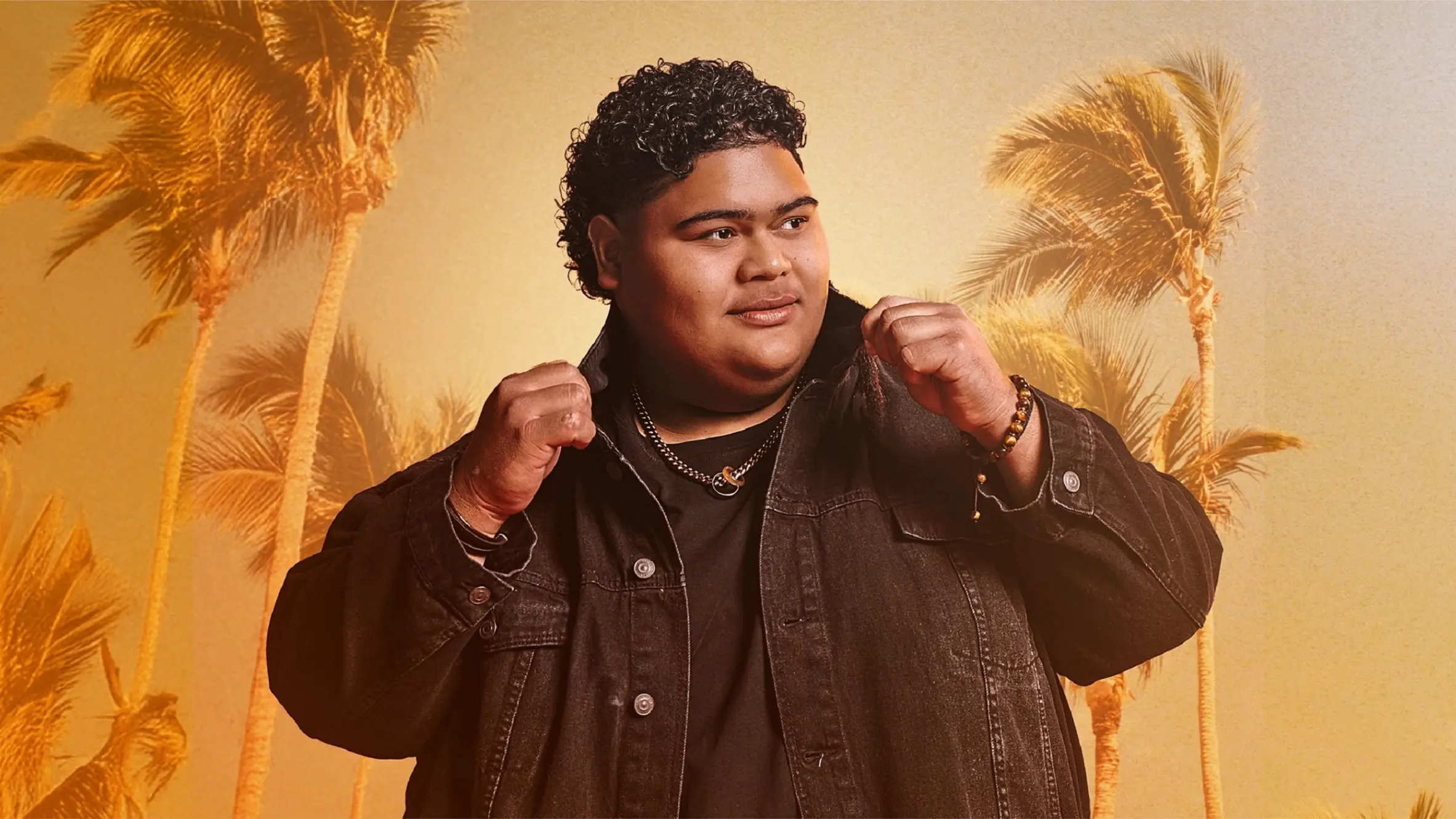 Person with curly hair wearing a black jacket and necklace, posing with fists raised against a background of palm trees.