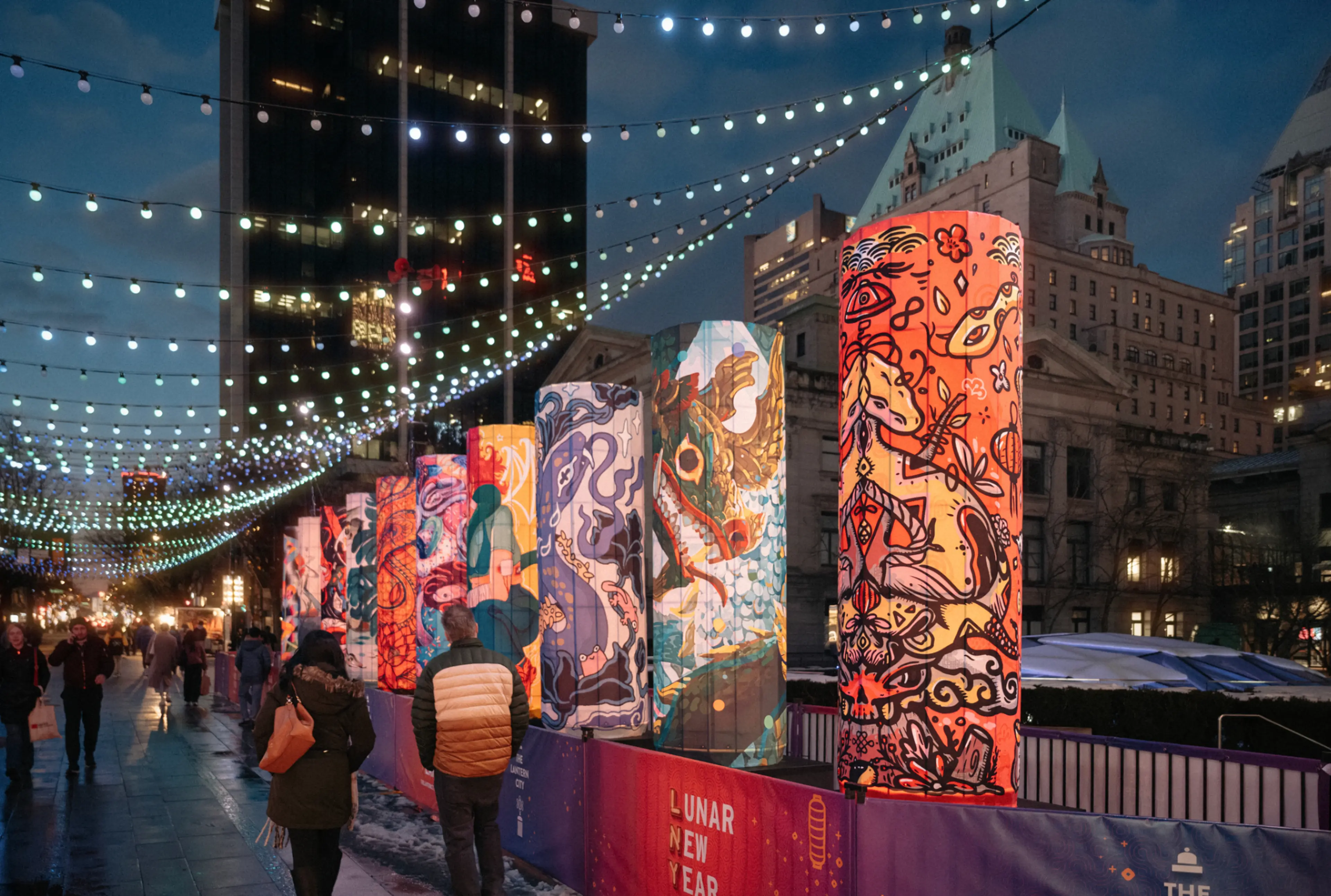 Colorful cylindrical lanterns with artistic designs lit up along a city street at night with string lights overhead and people walking.