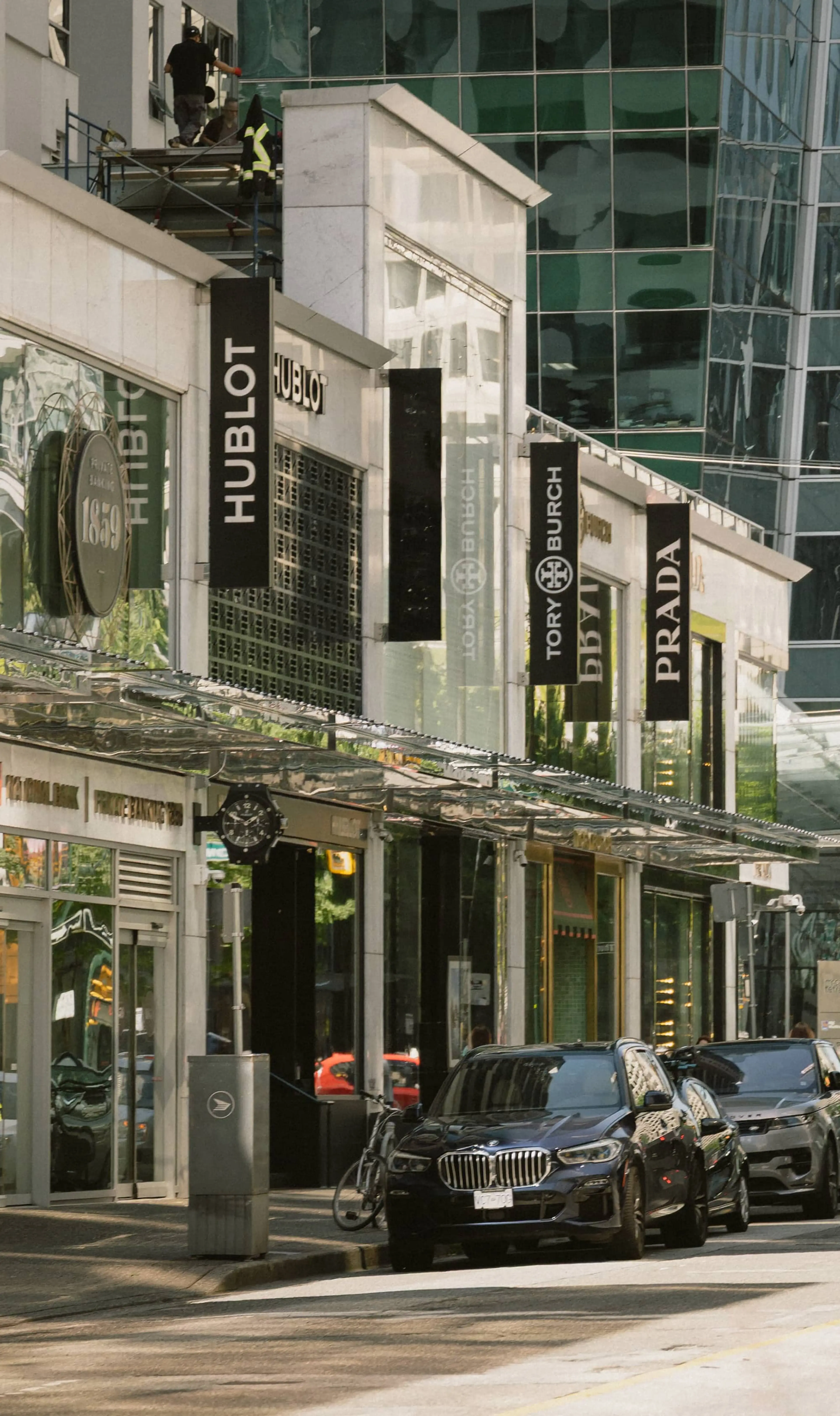 Street view of luxury brand stores with signs for Hublot, Tory Burch, and Prada, and parked cars in front.