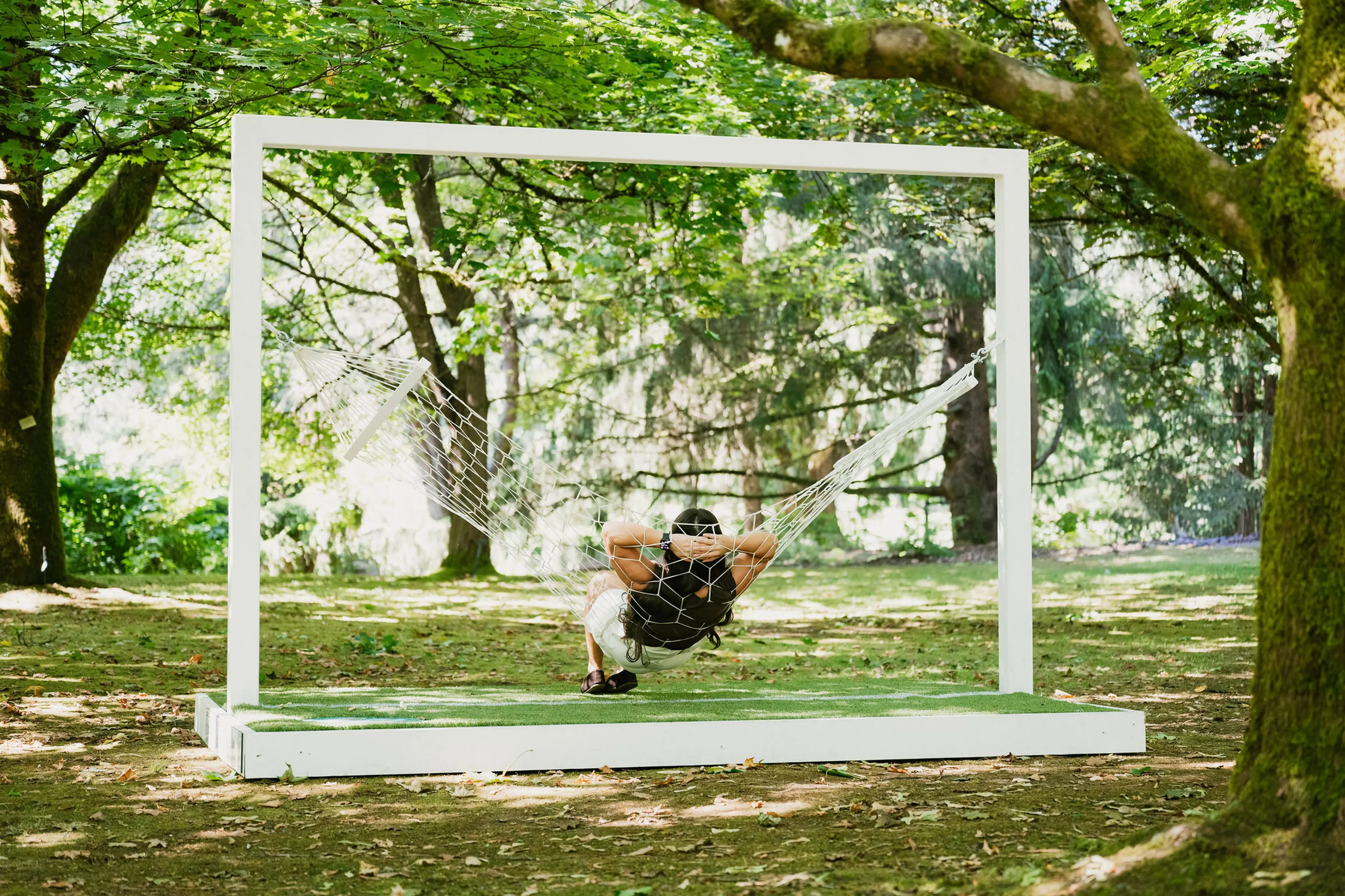 A person relaxing in a white hammock suspended within a large white rectangular frame in a shaded forest area.