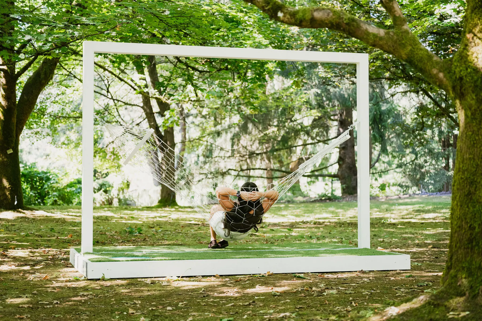 A person relaxing in a white hammock suspended within a large white rectangular frame in a shaded forest area.