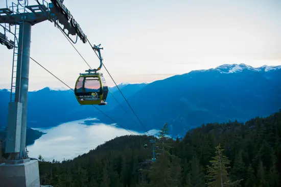 Gemini said A Sea to Sky Gondola cabin, featuring a yellow and black exterior, glides along a cable above a vast mountainous landscape during twilight. Below, a thick blanket of clouds or fog fills the valley over the water, while silhouettes of evergreen trees cover the foreground hills. In the distance, rugged, snow-capped mountain peaks stand against a pale blue and soft pink sky, capturing the serene atmosphere of Squamish, British Columbia.