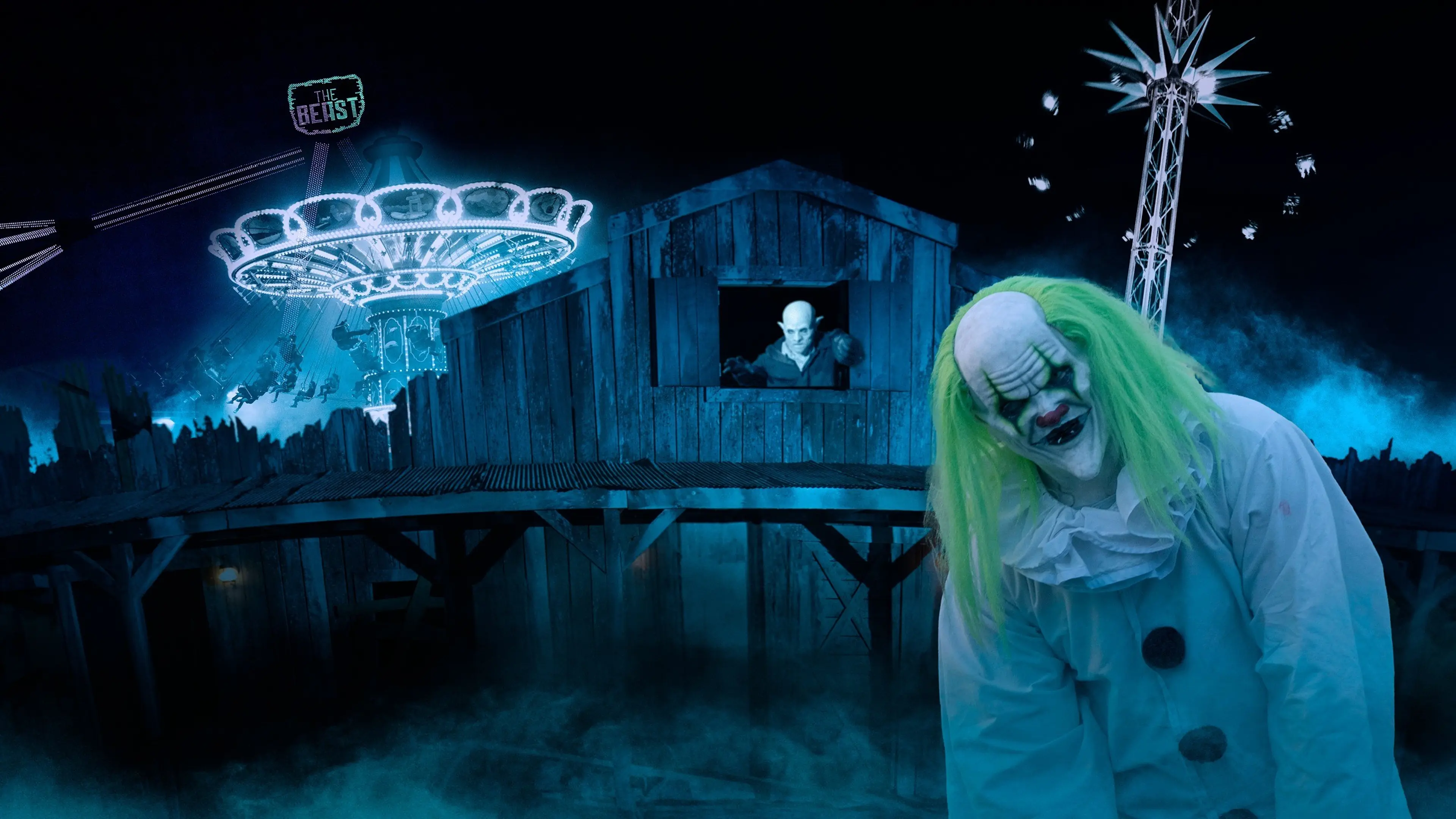 Clown with green hair and white costume leaning forward in front of a wooden haunted house with a vampire figure in the window and illuminated amusement rides in the background.