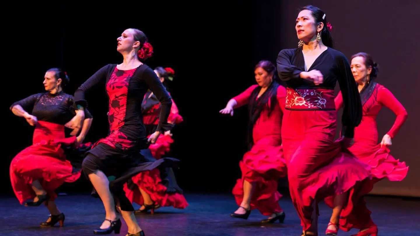 Six dancers performing flamenco in red and black dresses with flowers in their hair on stage.