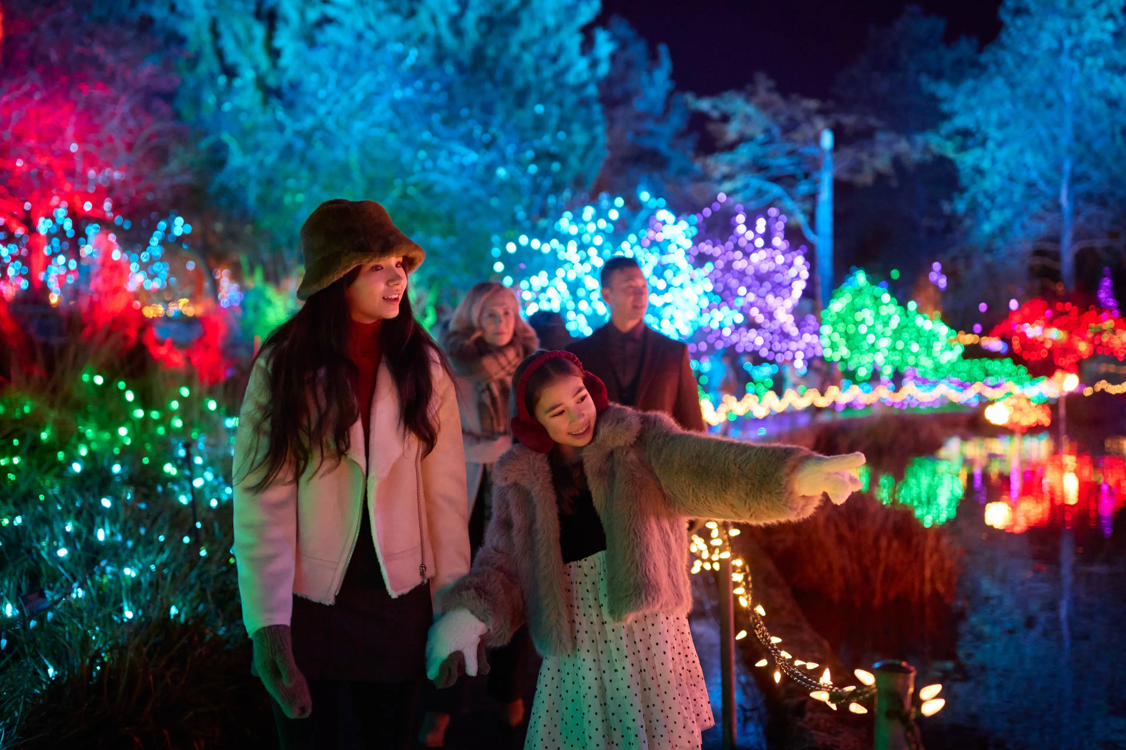 A family of four strolls along a decorated pond at VanDusen Garden’s Festival of Lights. The two daughters walk ahead, with the younger one pointing toward the glowing light displays that sparkle around them at dusk.