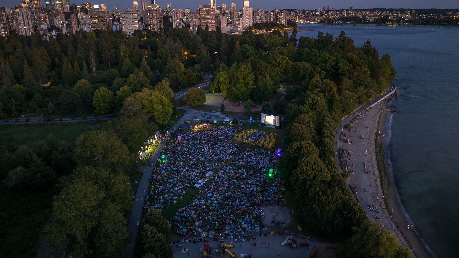 A large crowd gathered on a grassy area in a park at dusk watching an outdoor movie on a big screen, with a city skyline and water in the background.