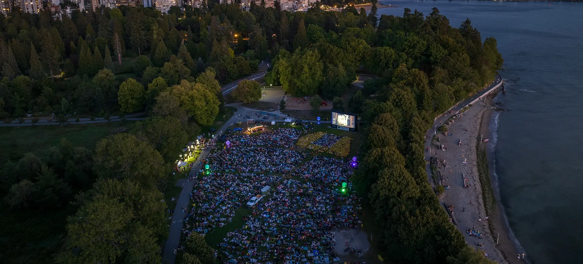 A large crowd gathered on a grassy area in a park at dusk watching an outdoor movie on a big screen, with a city skyline and water in the background.