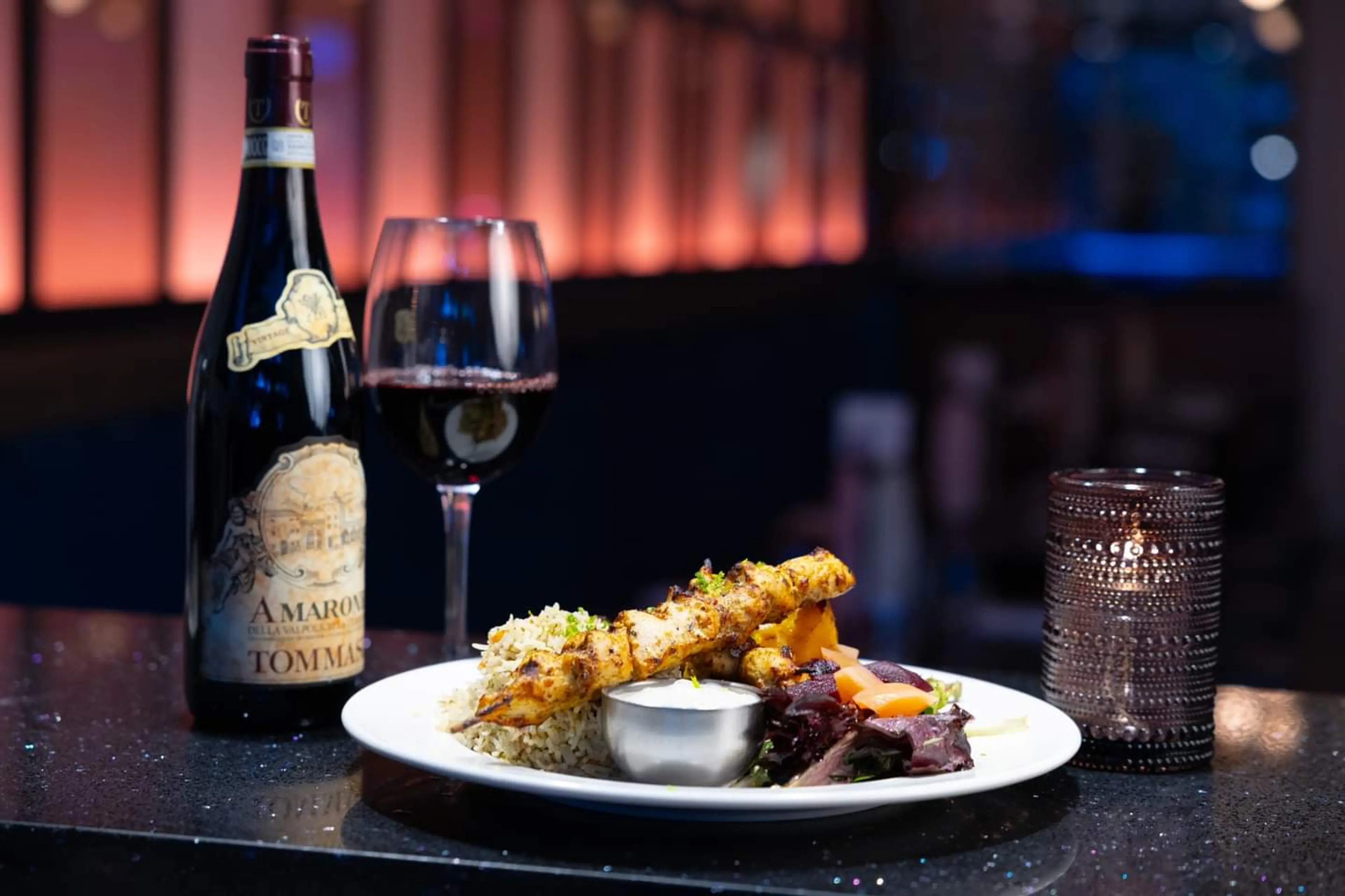 Plate with grilled chicken skewers, rice, salad, and sauce next to a bottle and glass of red wine on a dark table with a lit candle at the Four Olives restaurant in Vancouver.