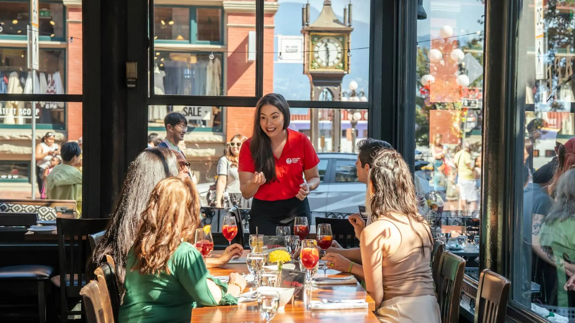 A waitress in a red shirt talks to four seated women at a restaurant table with drinks and menus, with a street clock visible outside.