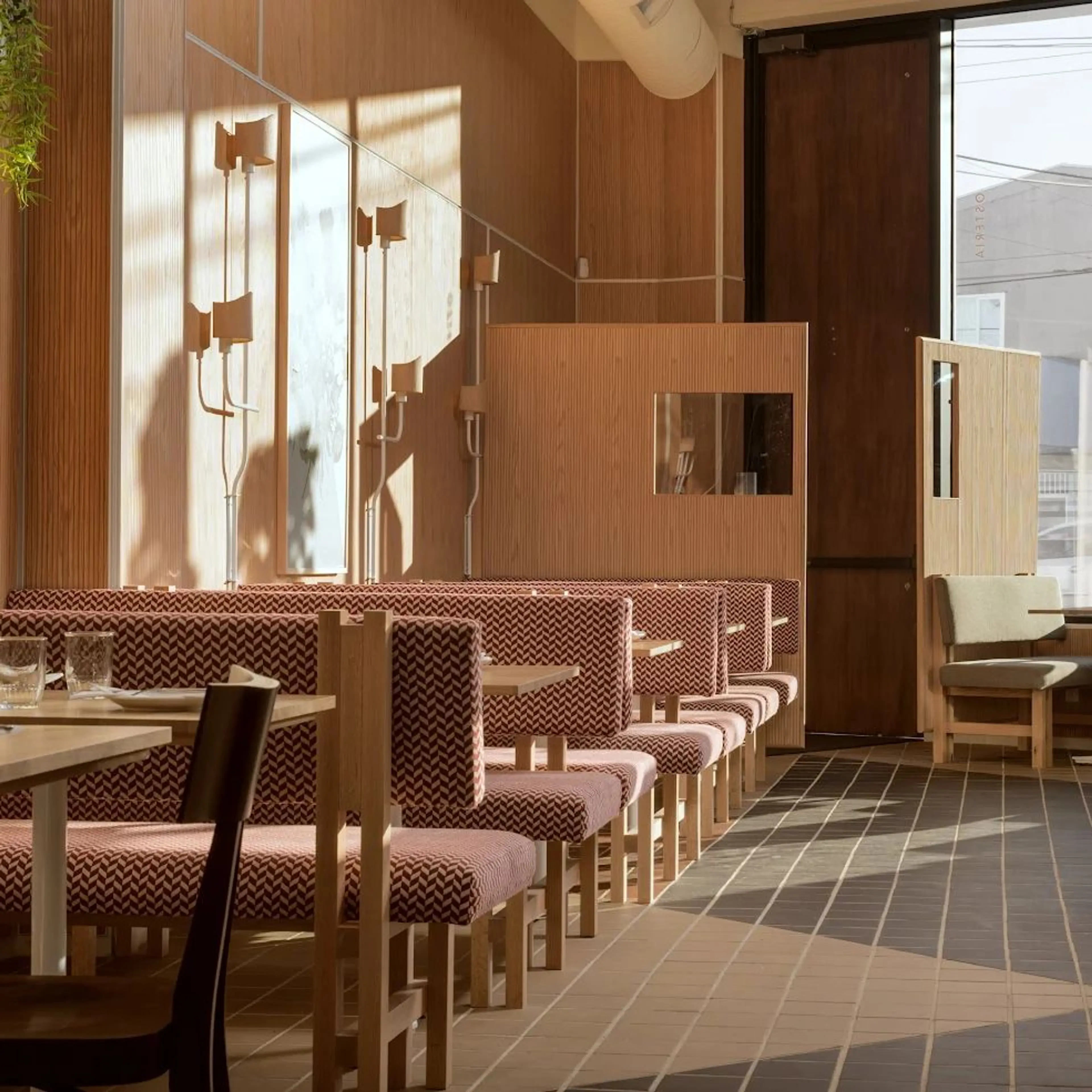 Sunlit restaurant interior with patterned booth seating, wooden tables, and a large window casting shadows.
