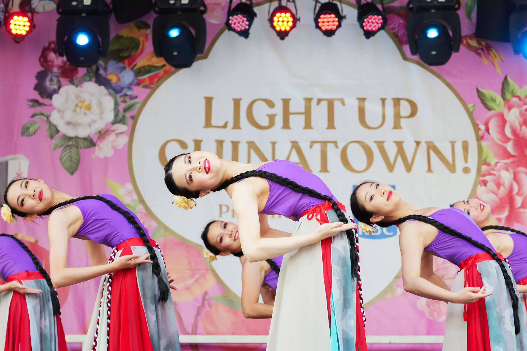 Five dancers in purple tops and colorful skirts performing a synchronized backbend on stage with a floral backdrop reading Light Up Chinatown.
