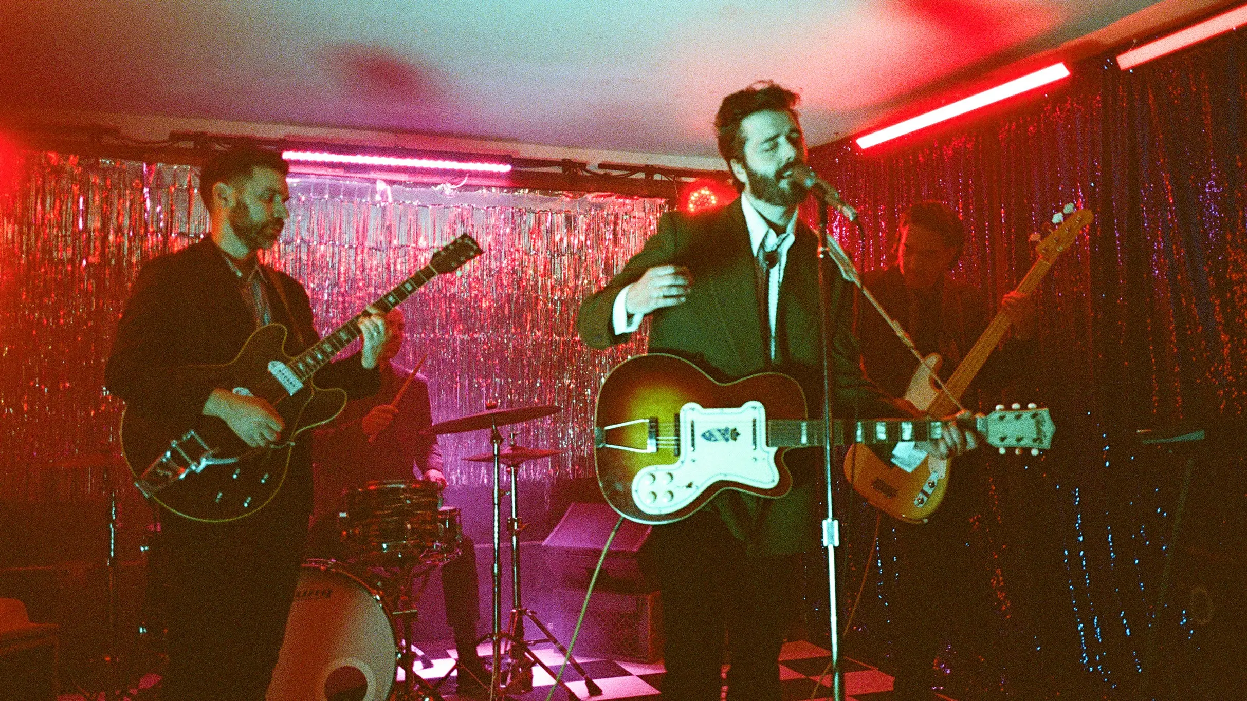 Four musicians performing on stage with guitars and drums under red and purple lighting with a shiny backdrop.
