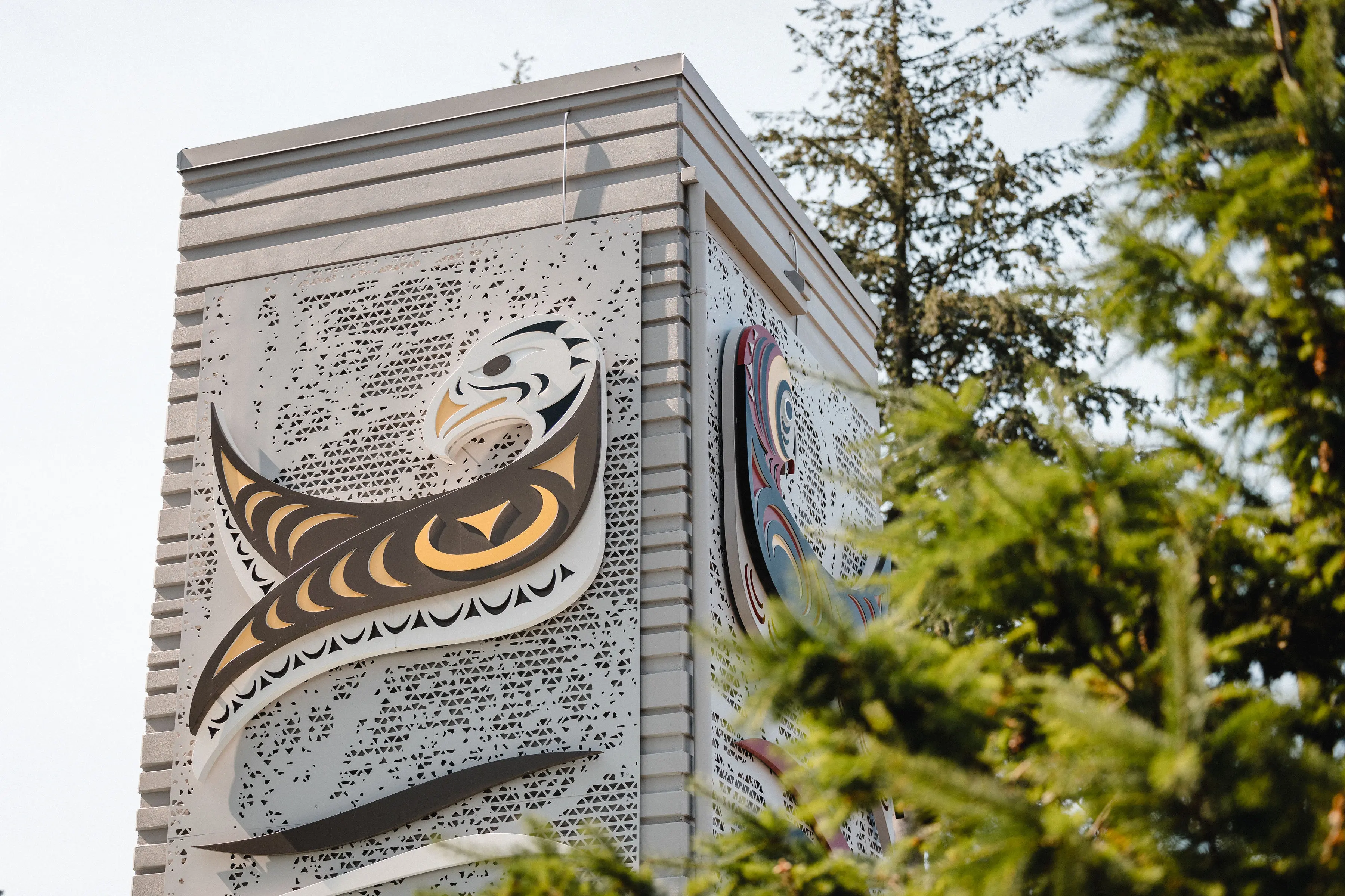 Corner of a building with Native American-style eagle artwork on metal panels, partially obscured by green trees.
