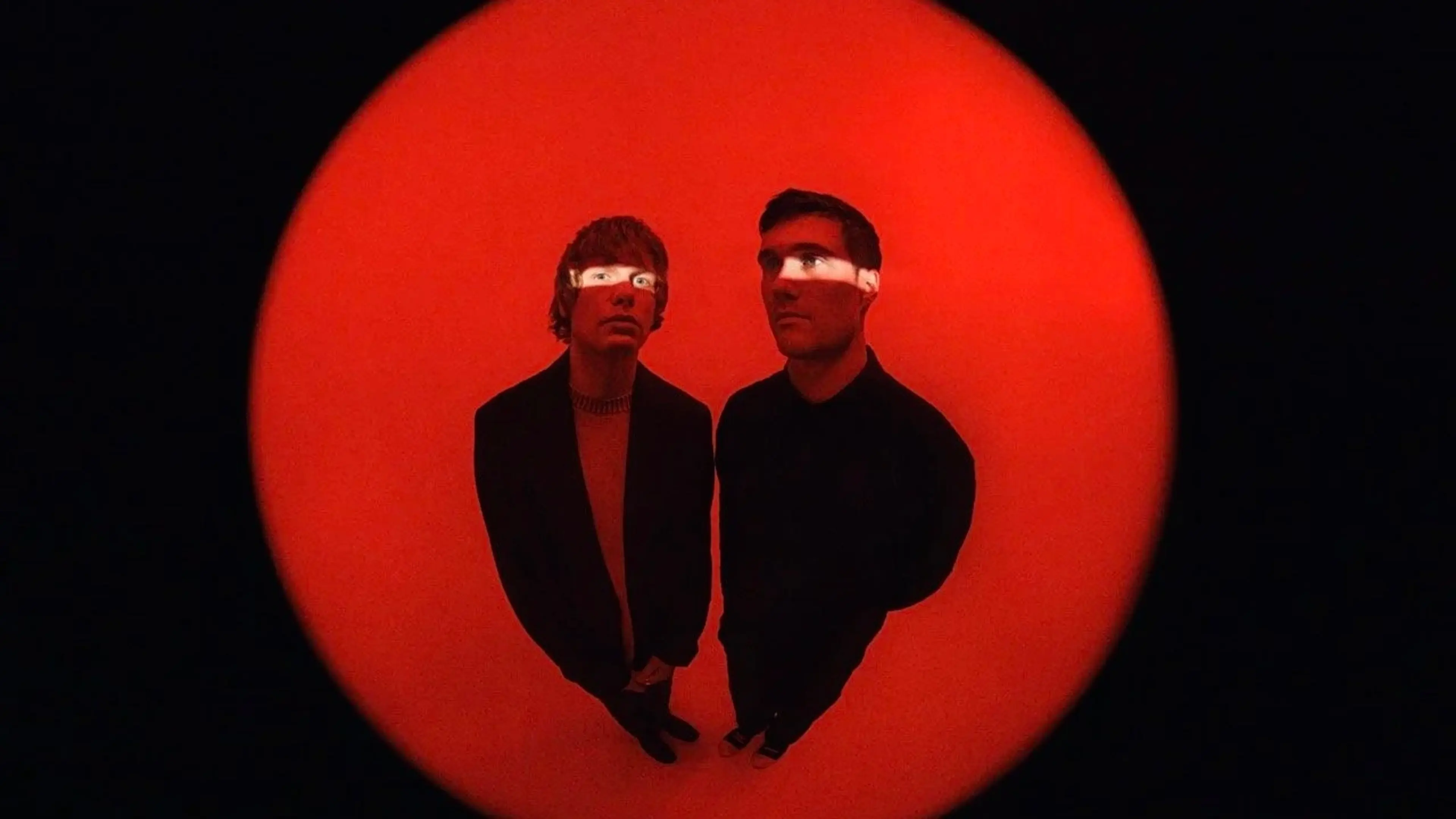 Two people standing on a red circular floor with a black background, light strip across their eyes.