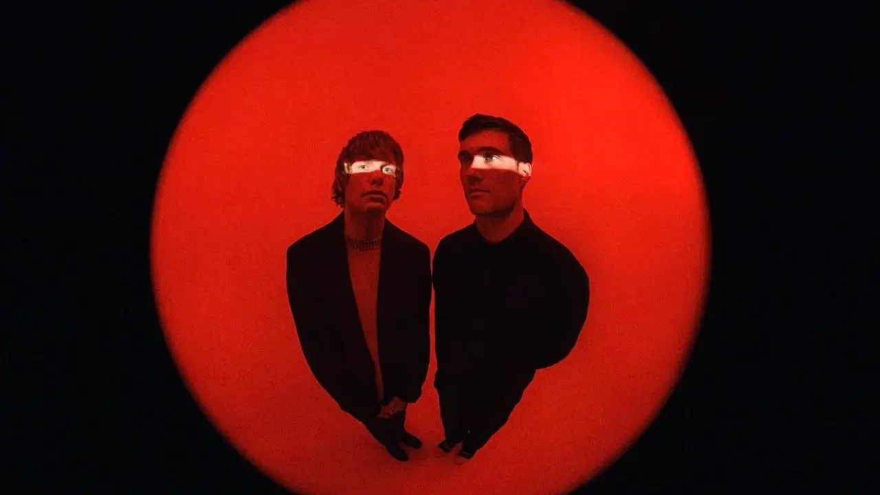 Two people standing on a red circular floor with a black background, light strip across their eyes.
