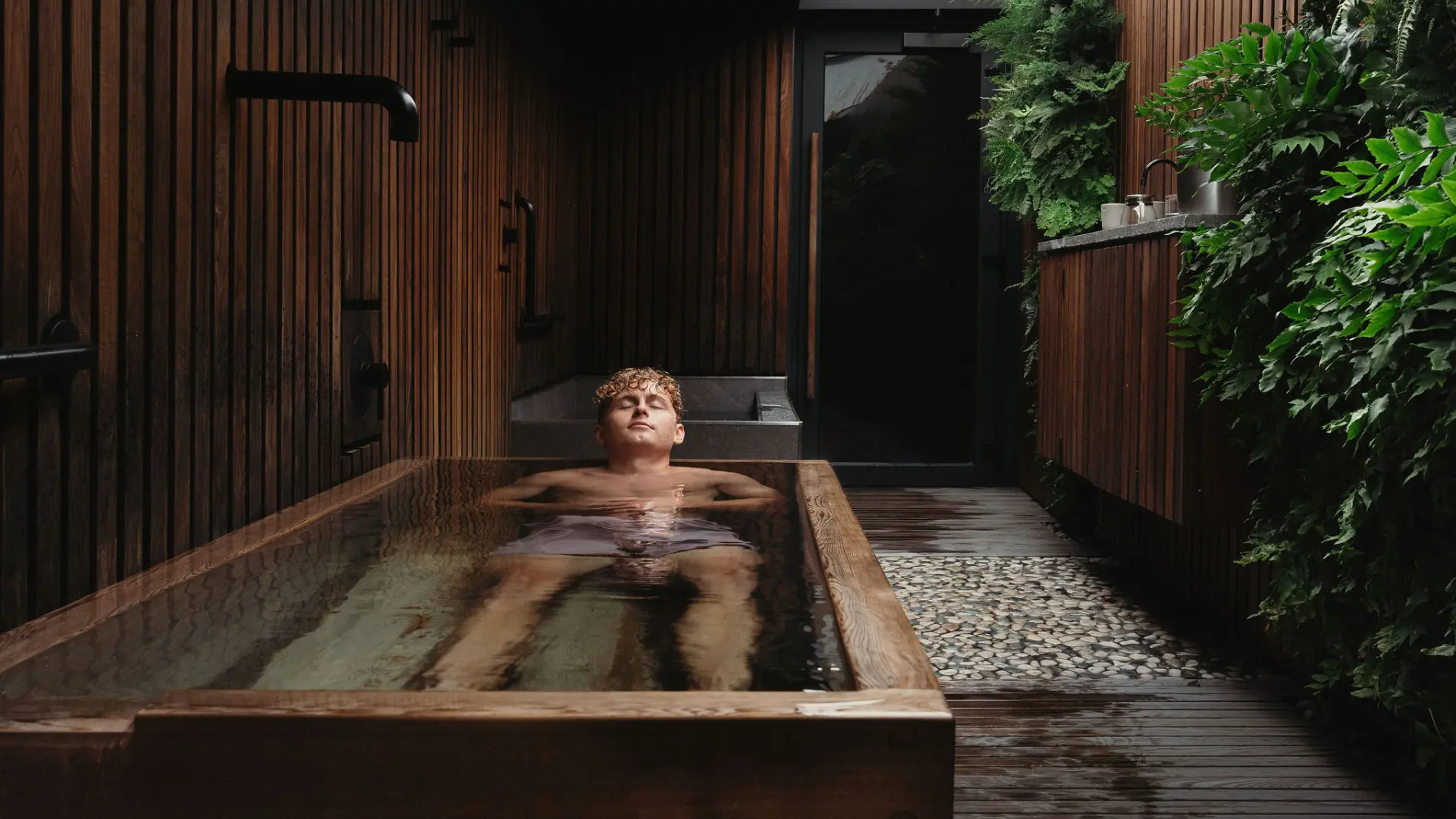 Person relaxing in a wooden bathtub in a spa-like room with wooden walls and green plants.