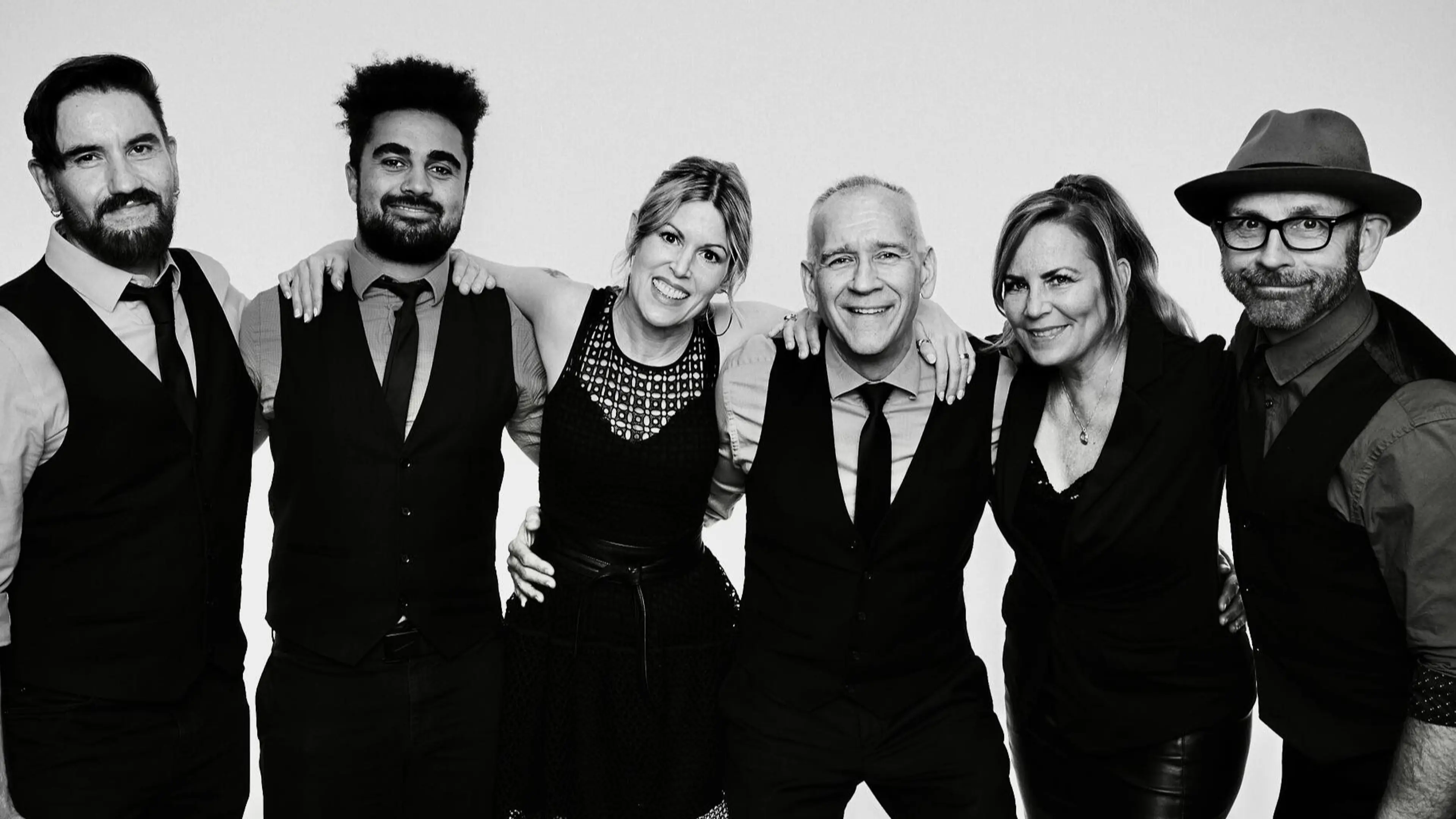 Group of six people standing close together, smiling, wearing formal black and white attire against a plain background.
