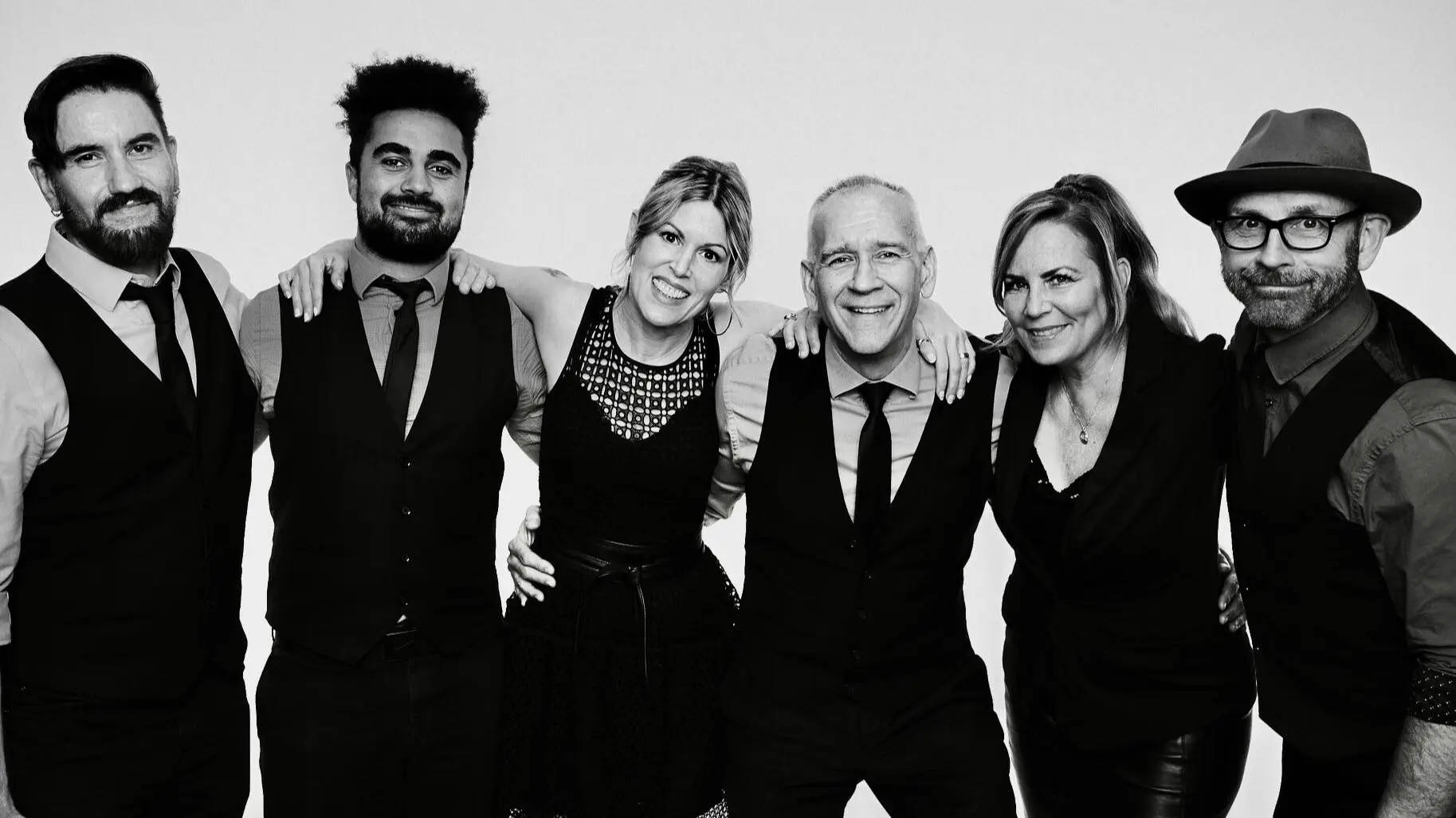 Group of six people standing close together, smiling, wearing formal black and white attire against a plain background.