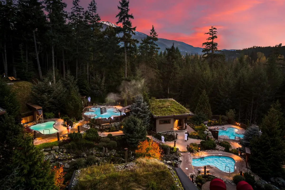 Outdoor spa with multiple hot tubs surrounded by trees and mountains under a pink sunset sky.
